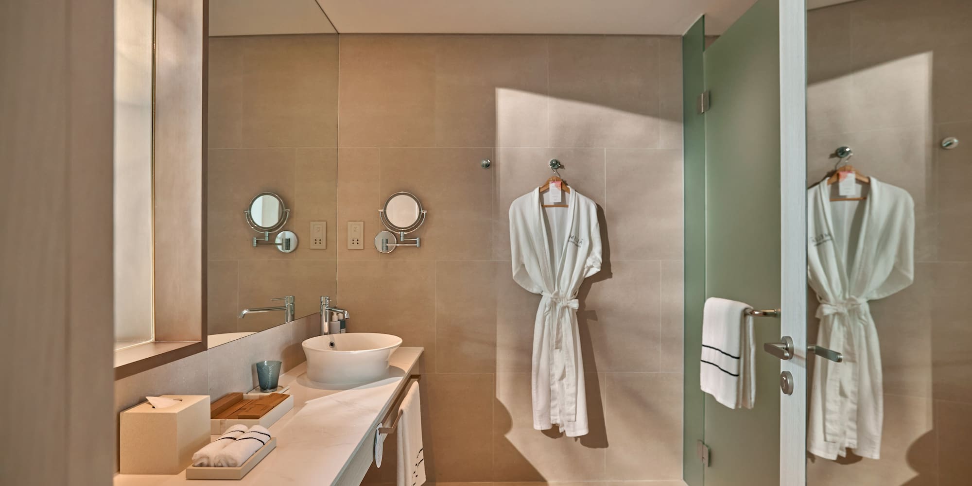 Modern hotel bathroom with white robe, vessel sink, and frosted glass shower.