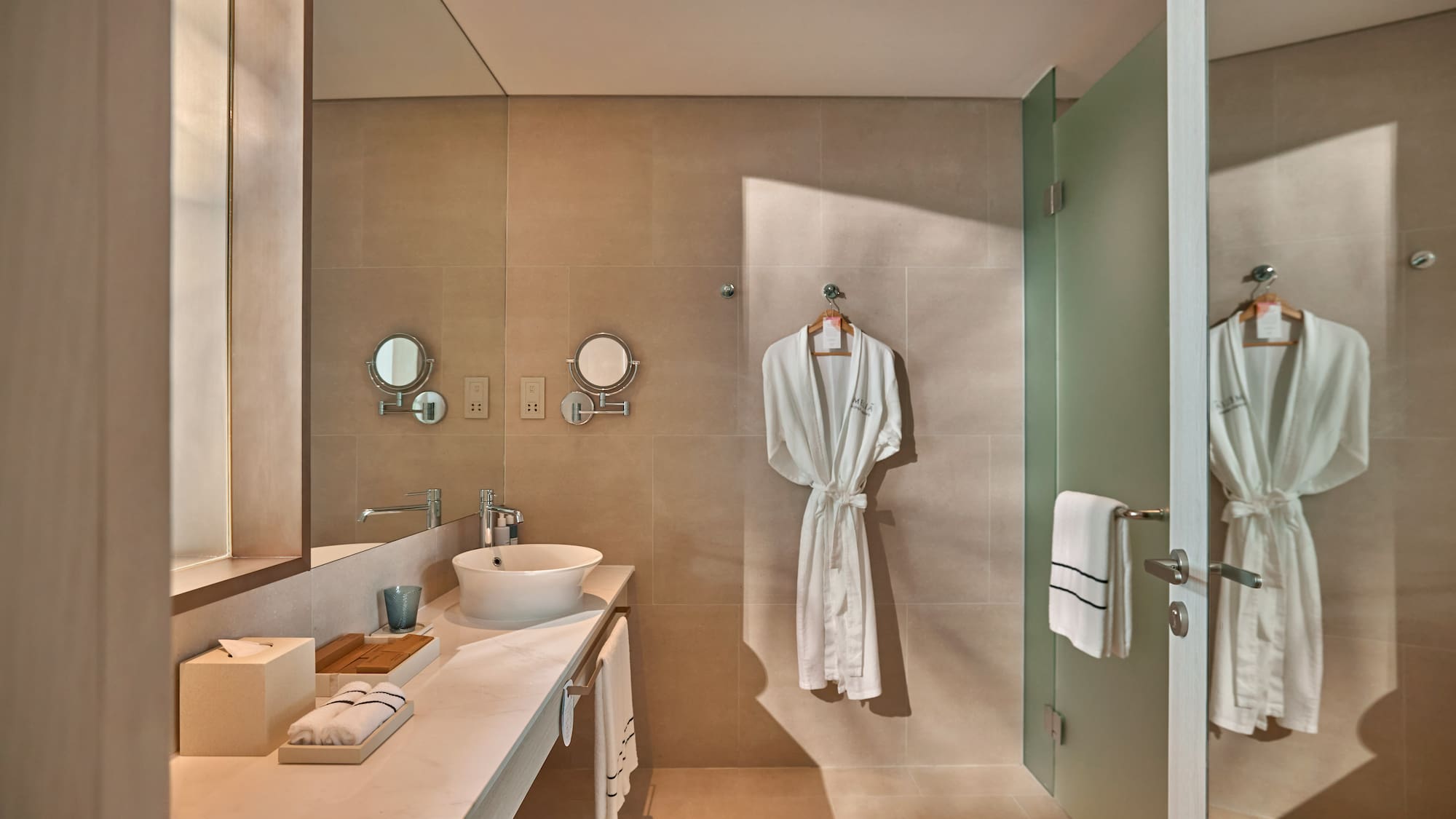 Modern hotel bathroom with white robe, vessel sink, and frosted glass shower.