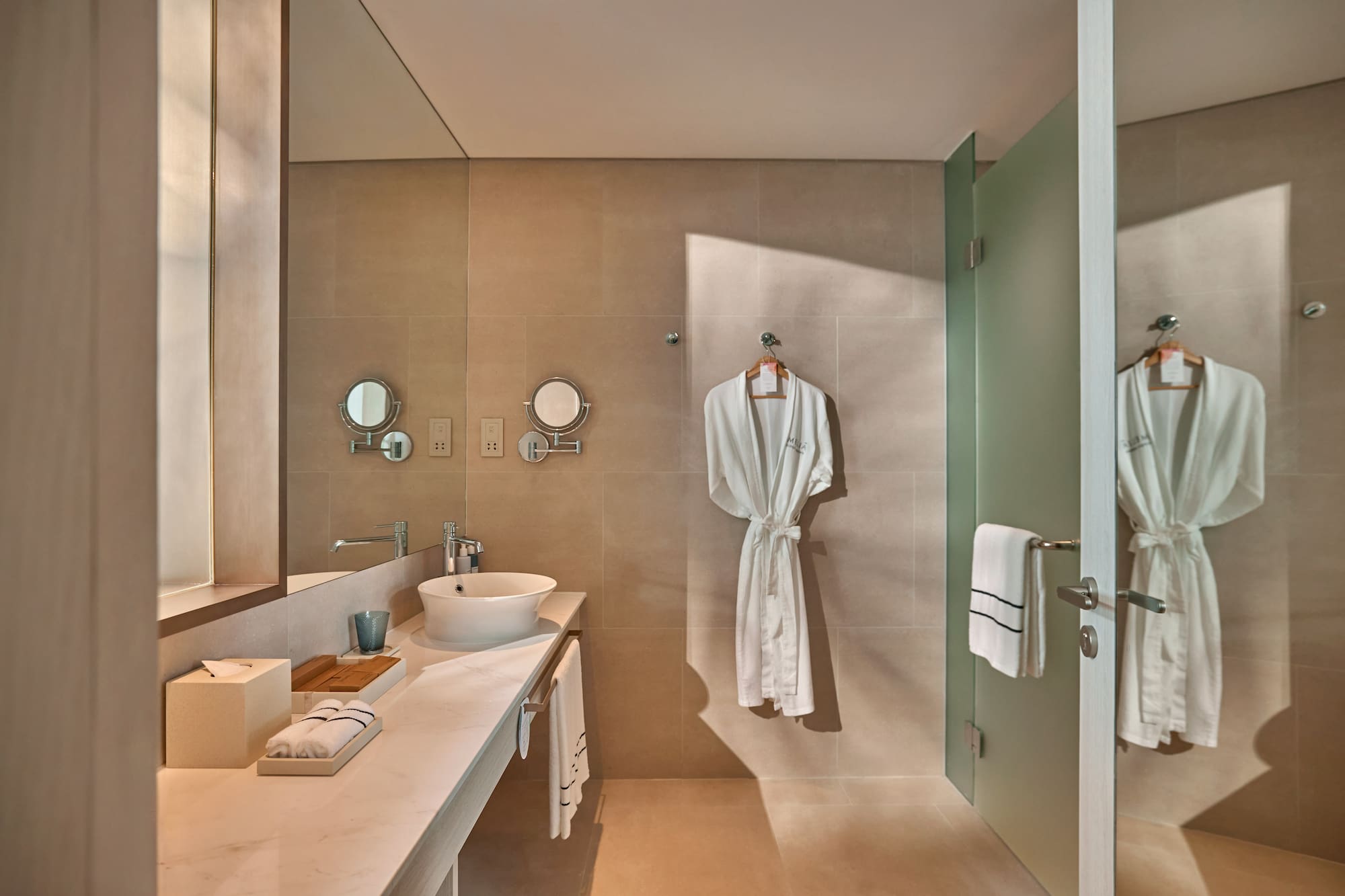 Modern hotel bathroom with white robe, vessel sink, and frosted glass shower.