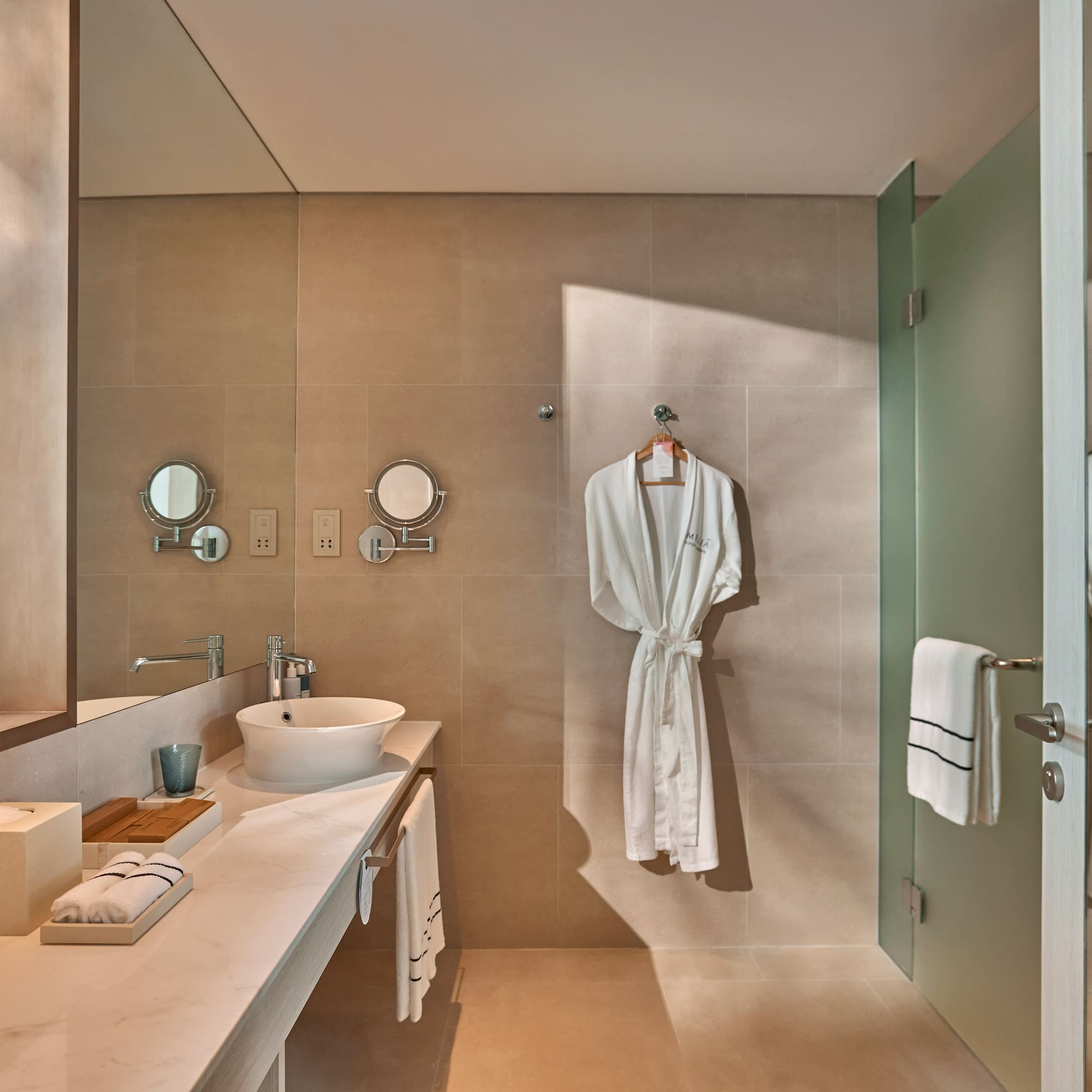 Modern hotel bathroom with white robe, vessel sink, and frosted glass shower.