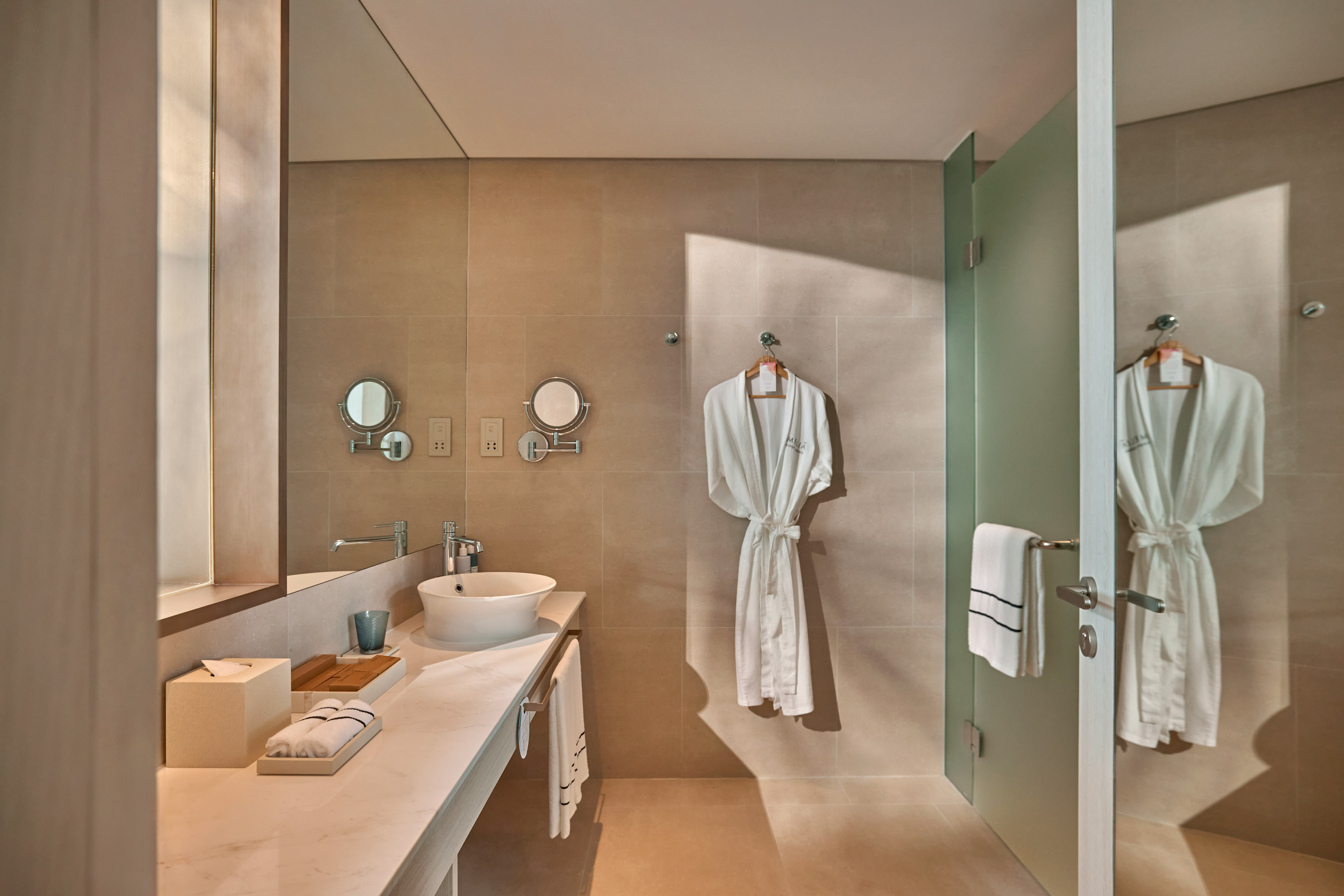 Modern hotel bathroom with white robe, vessel sink, and frosted glass shower.