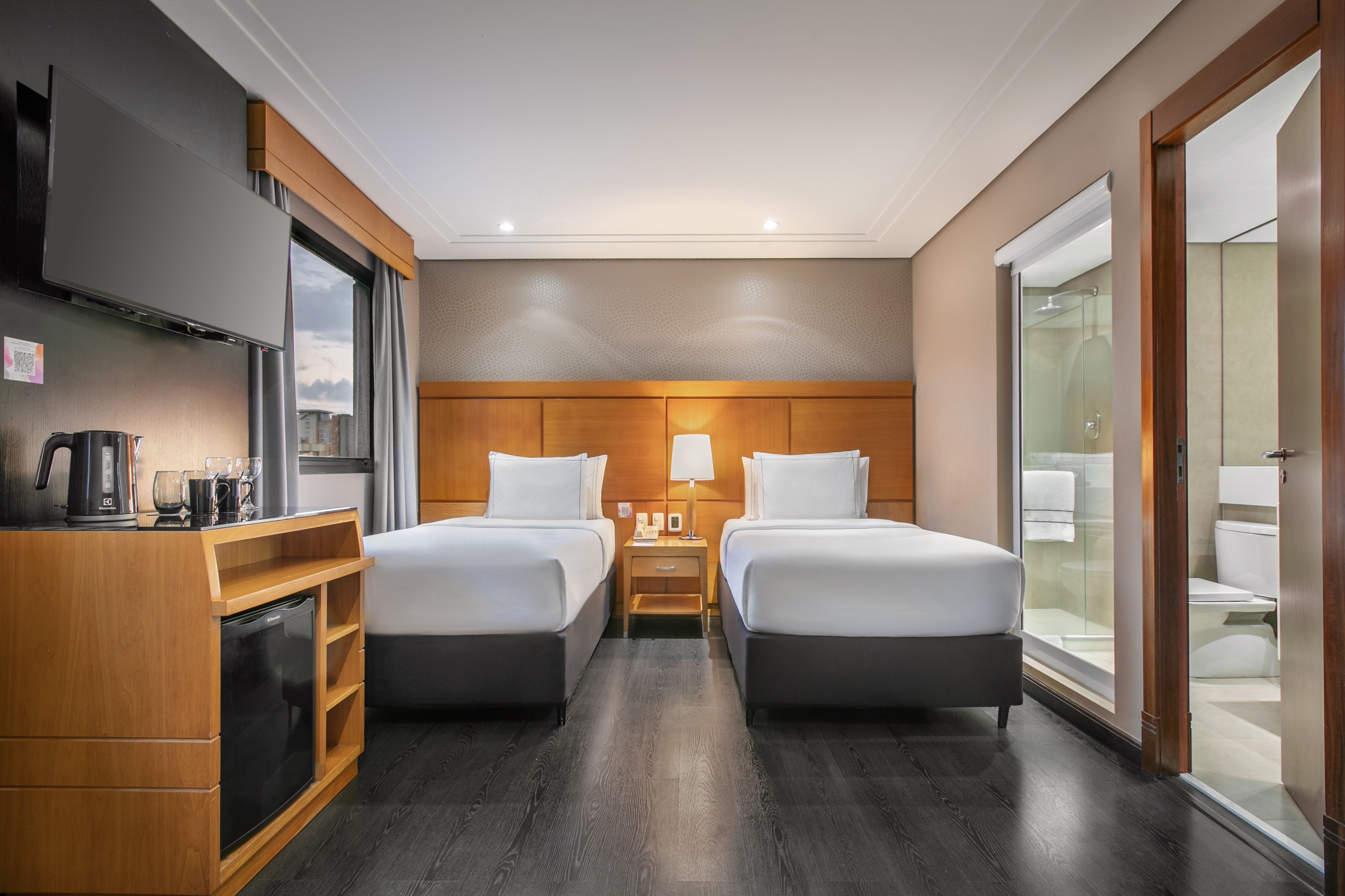 Modern hotel room with two twin beds, dark floor, TV, and mini-bar.