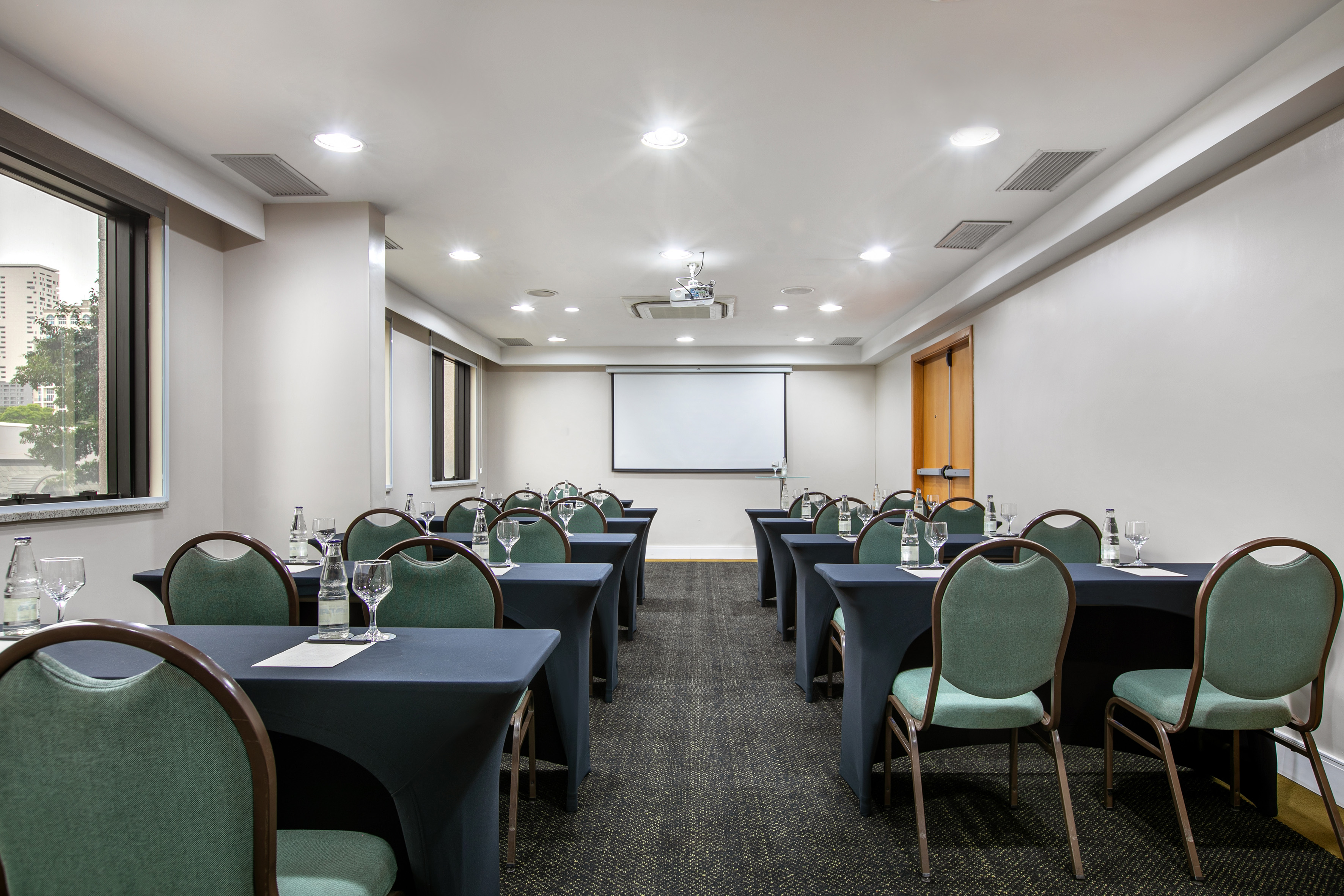 Conference room setup with projector screen, rows of dark tables, green chairs.
