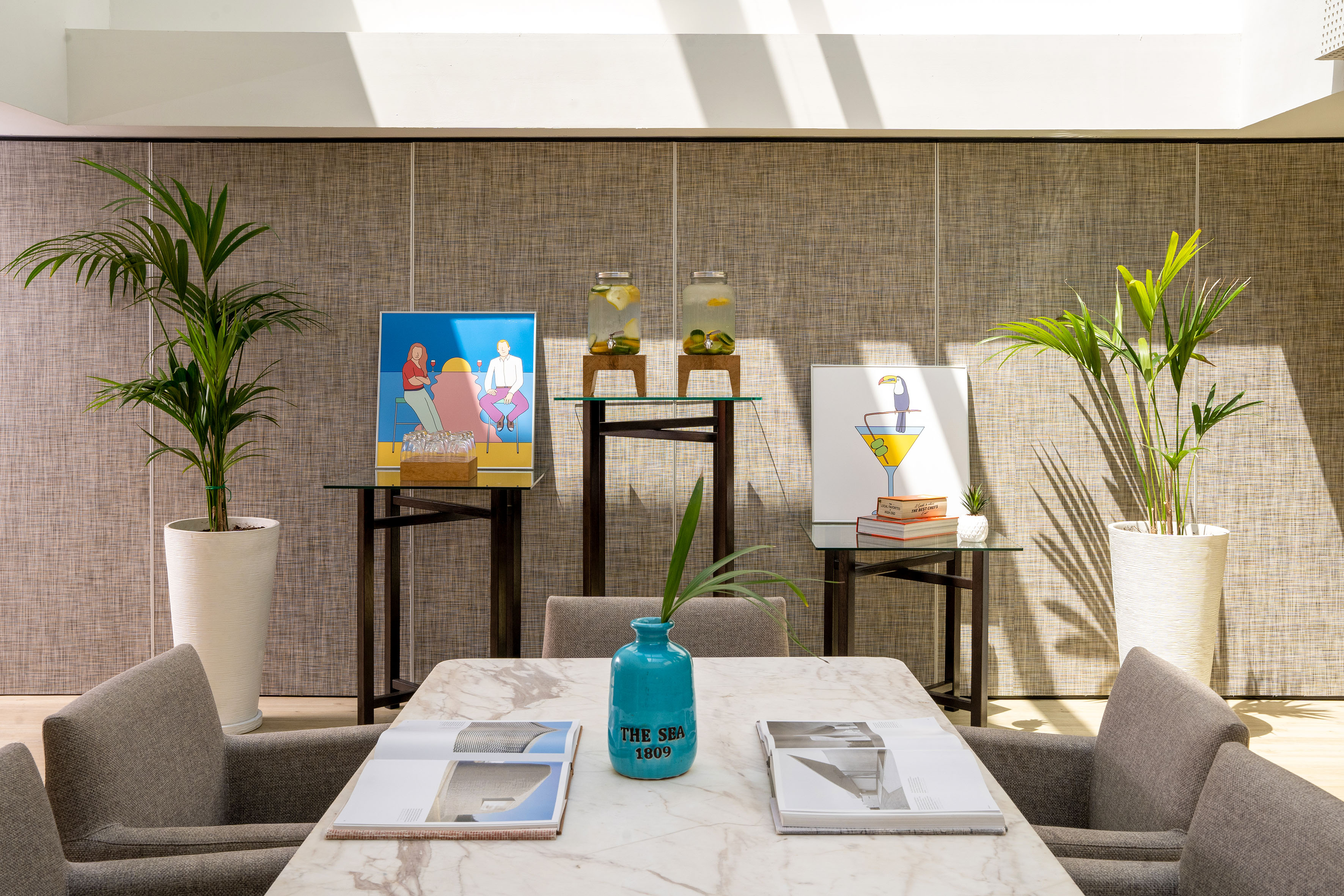 Modern lounge with plants, fruit water, marble table, and vibrant artwork.