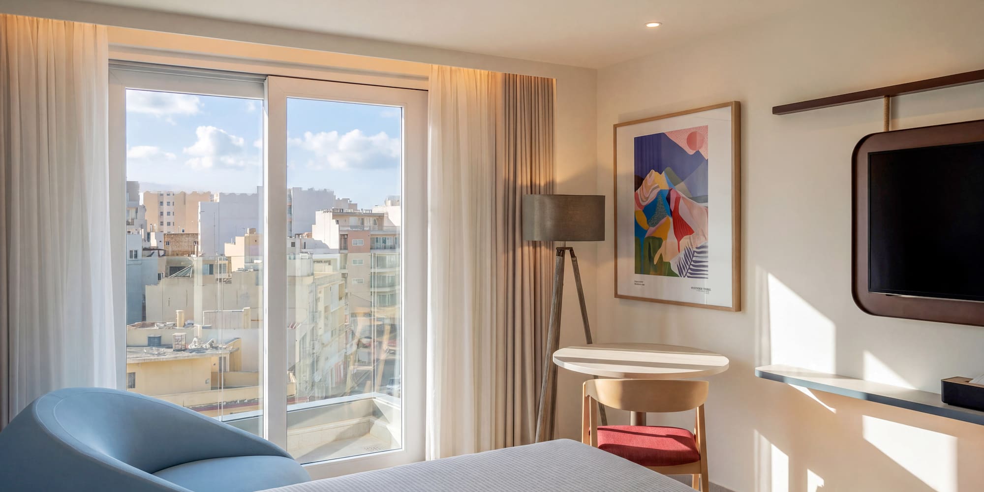 Modern hotel bedroom with city view, large window, abstract art, blue armchair.
