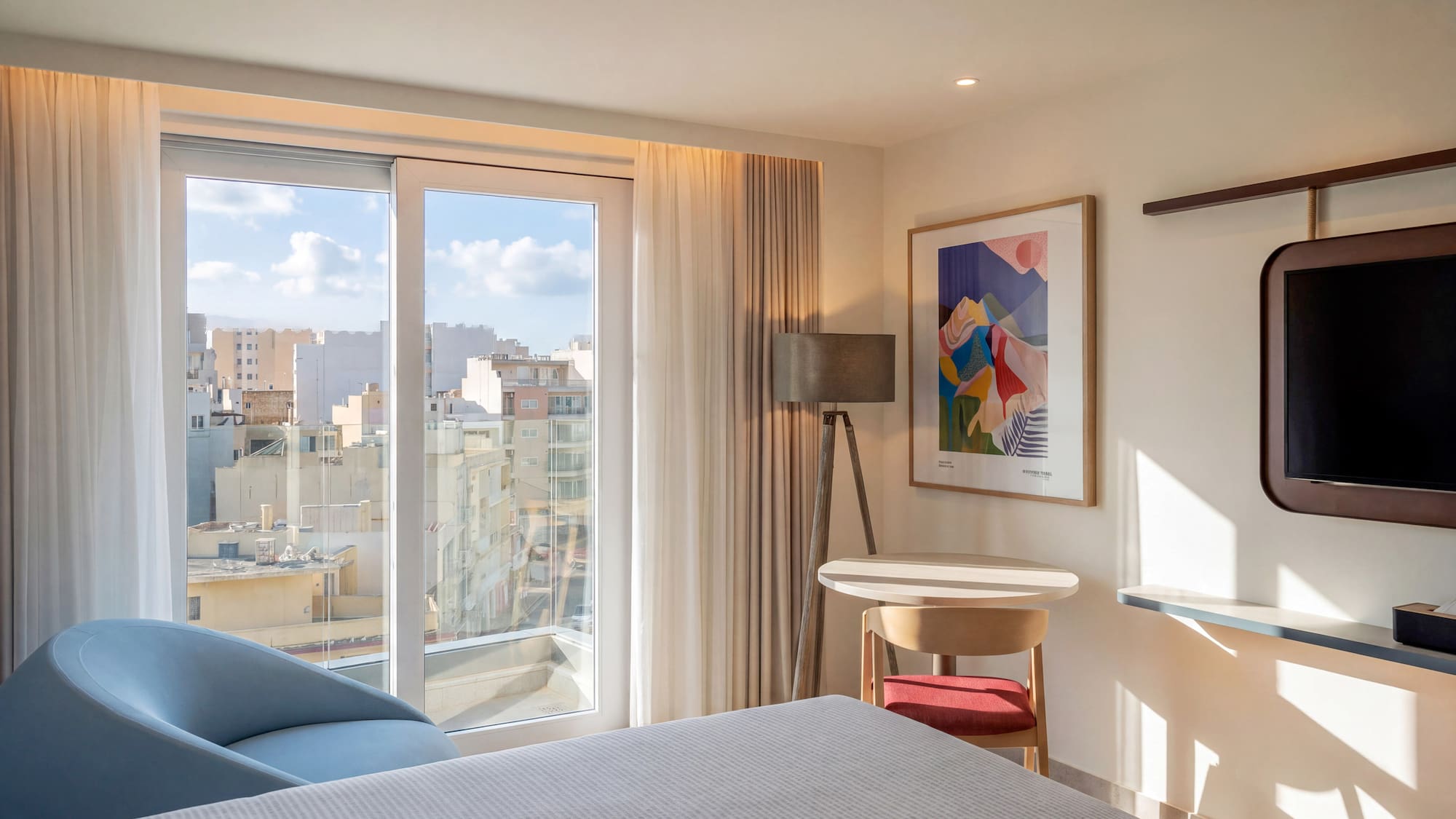 Bright hotel bedroom with expansive city view, modern furnishings.