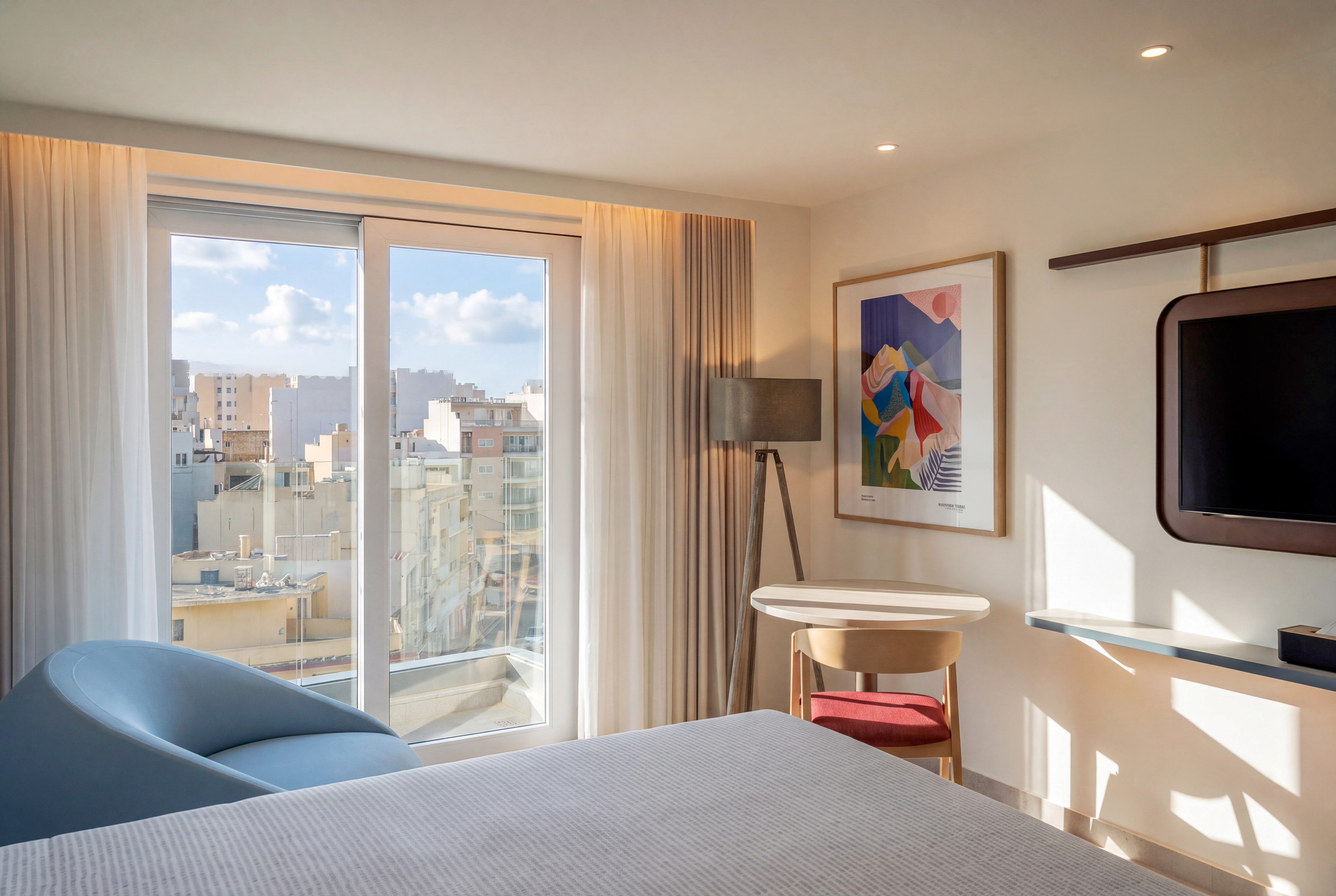 Modern hotel bedroom with city view, large window, abstract art, blue armchair.