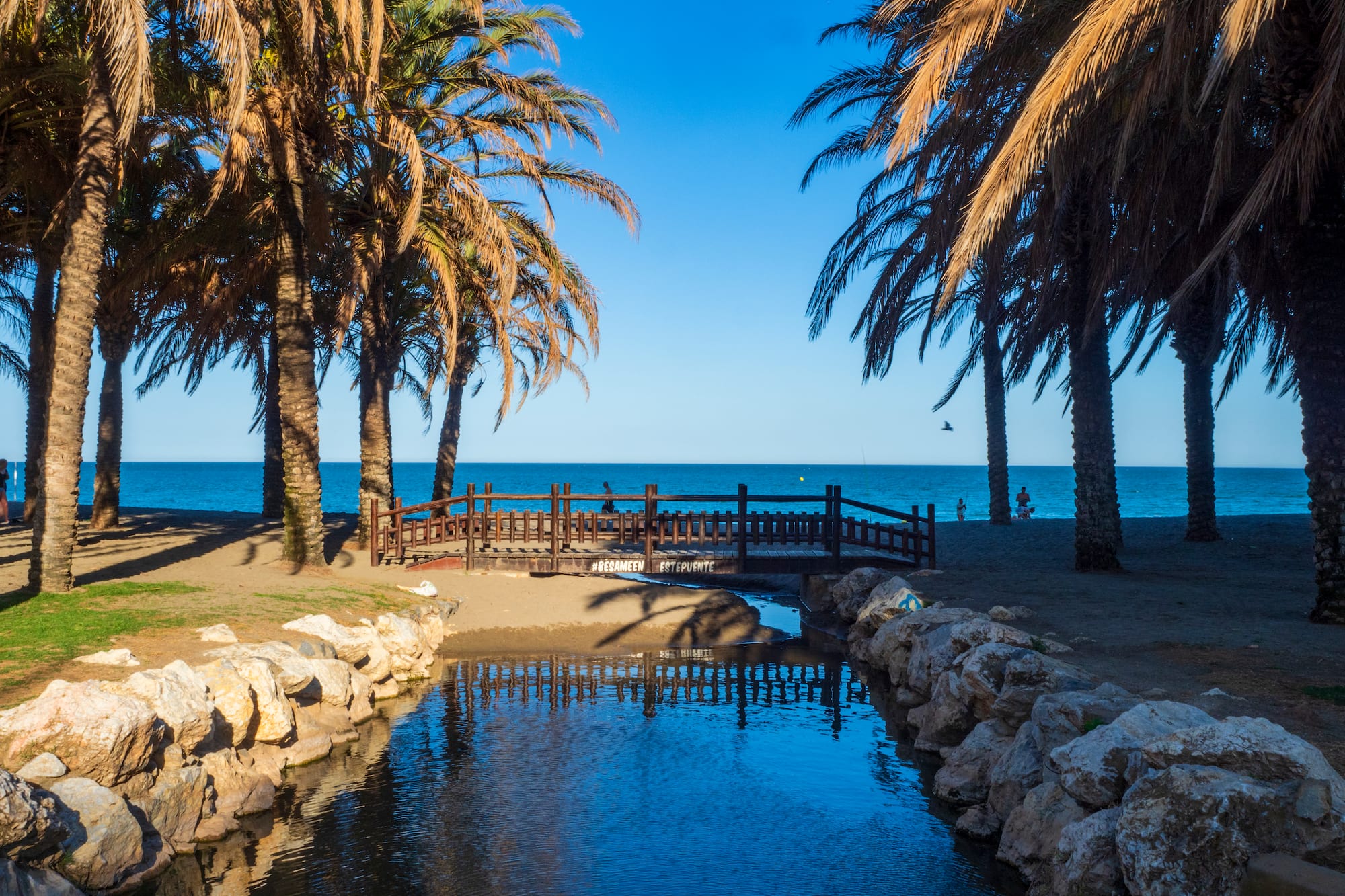 Tropical beach with palm trees, wooden bridge over canal, and blue sea.