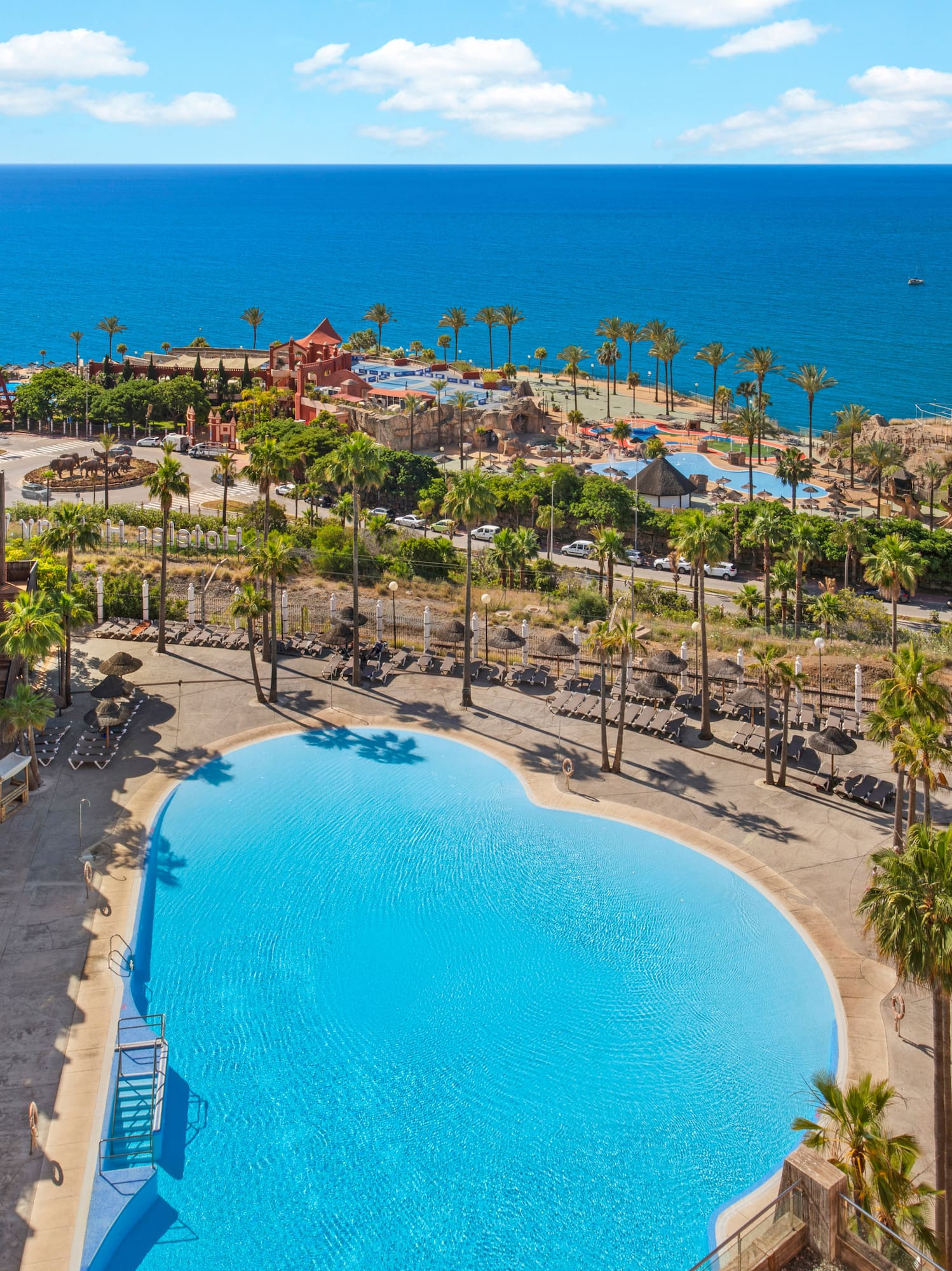 Tropical resort with large pool, palm trees, hotel, and ocean view.
