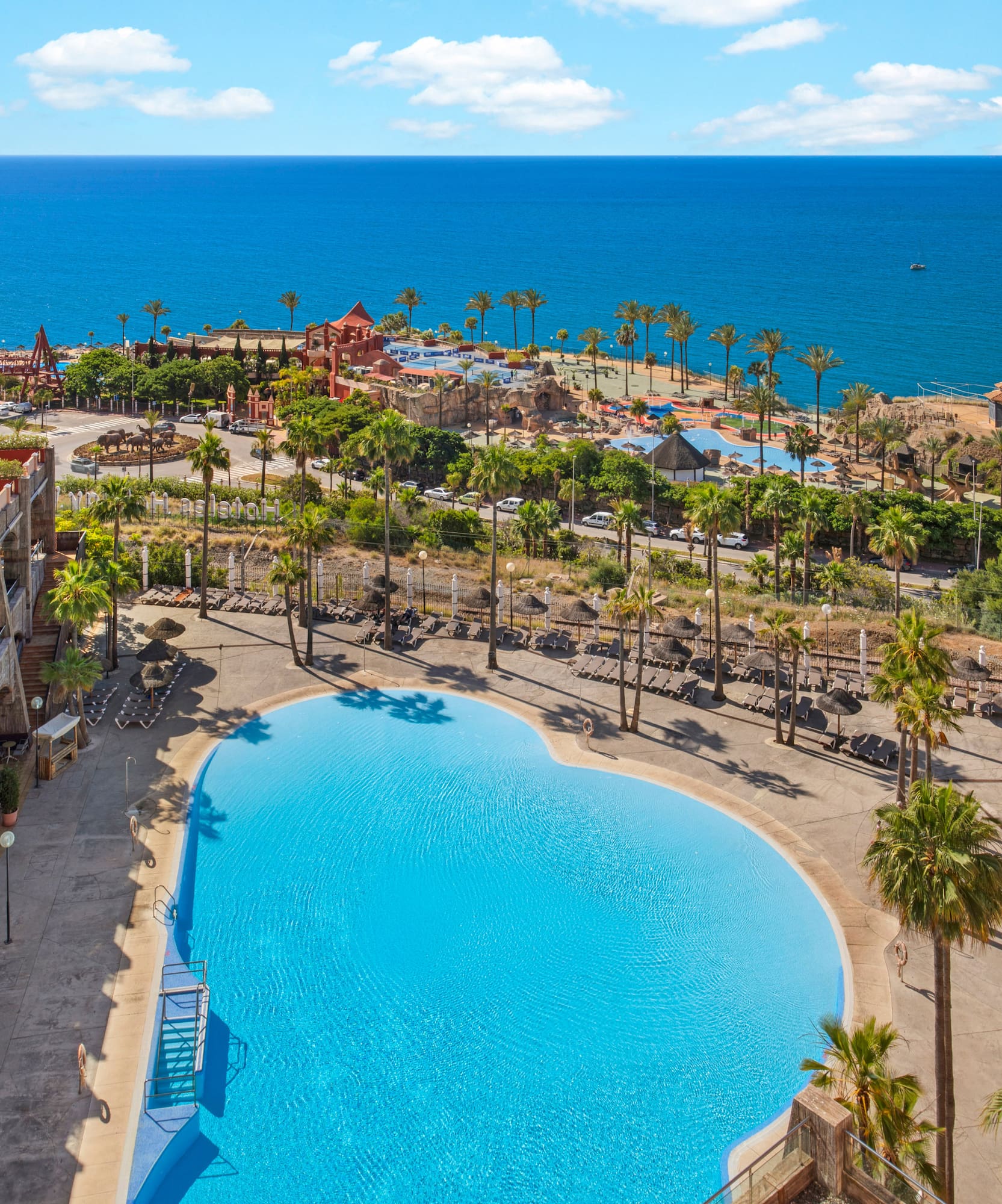 Tropical resort with large pool, palm trees, hotel, and ocean view.