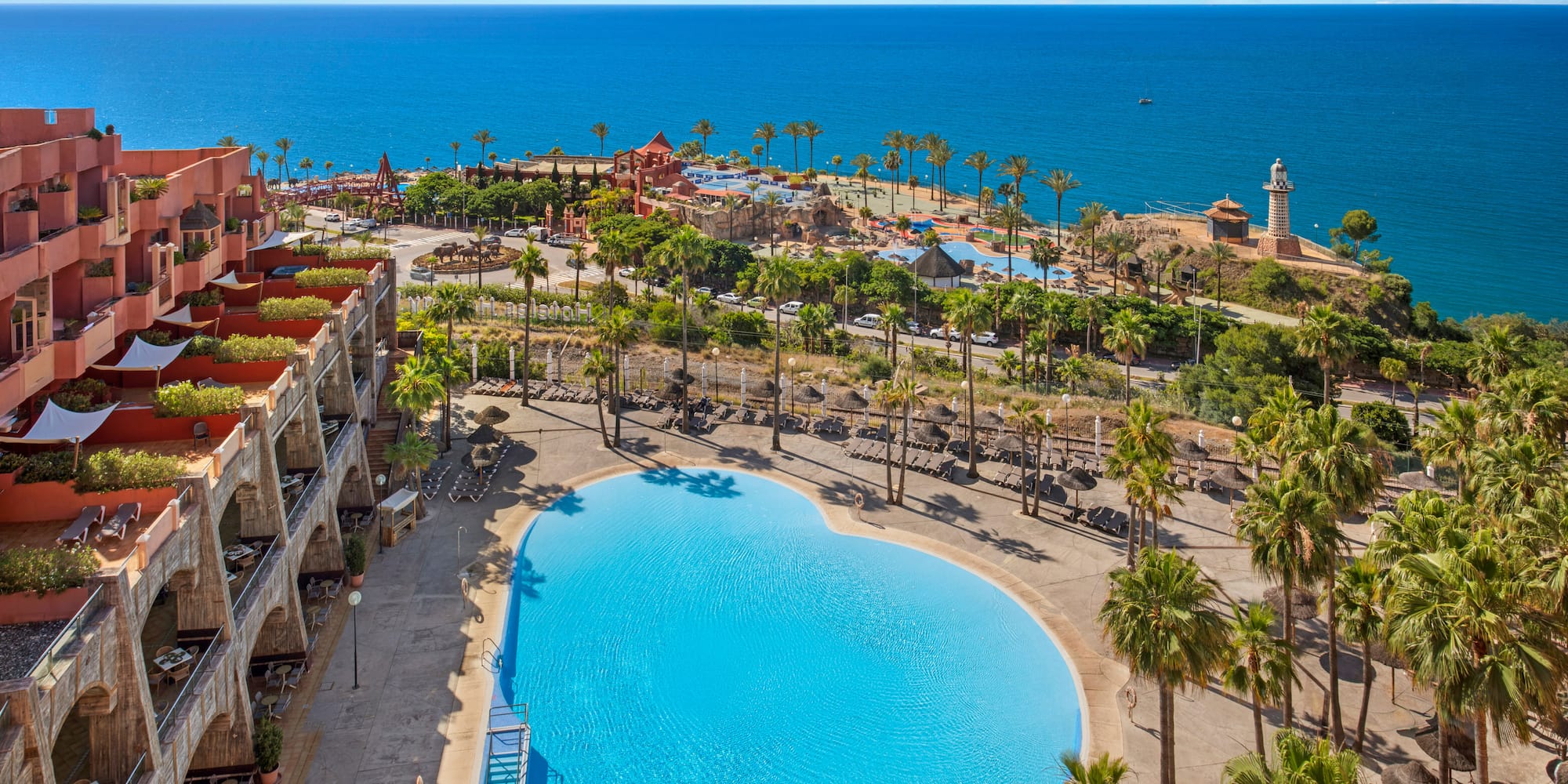Mediterranean resort with heart-shaped pool, palm trees, hotel, and ocean.