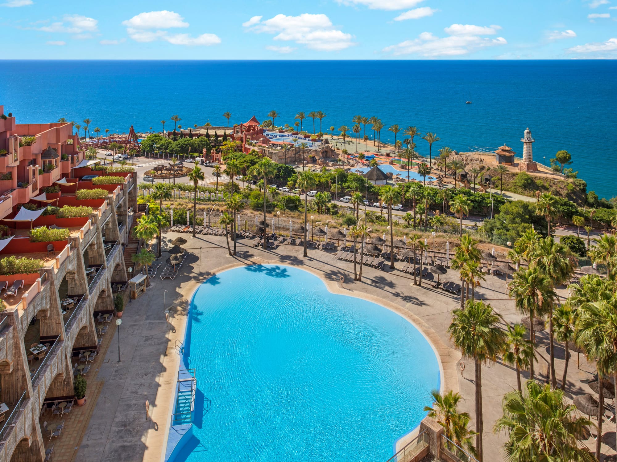 Mediterranean resort with heart-shaped pool, palm trees, hotel, and ocean.