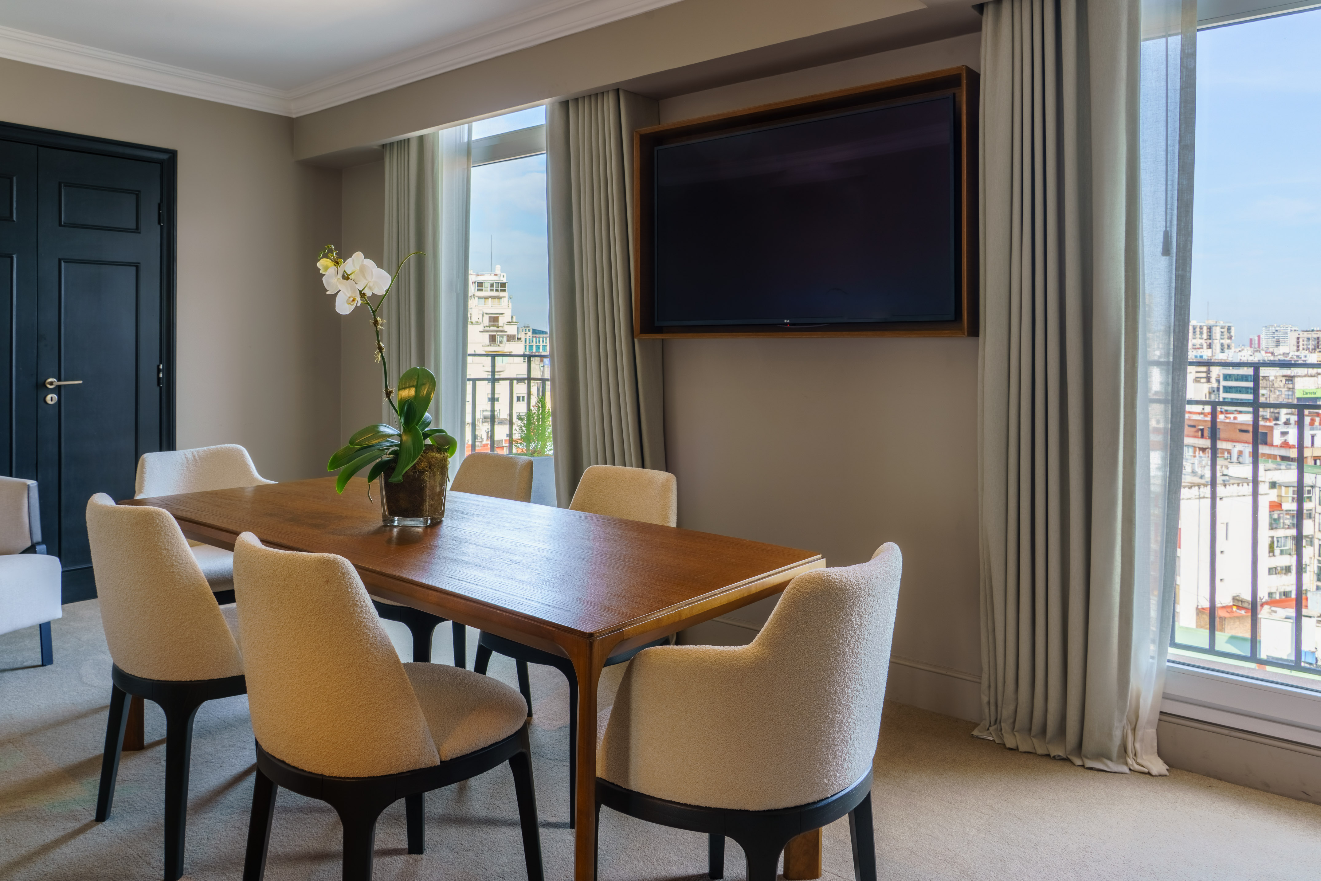 Modern hotel suite dining room with wood table, chairs, wall TV, city view.