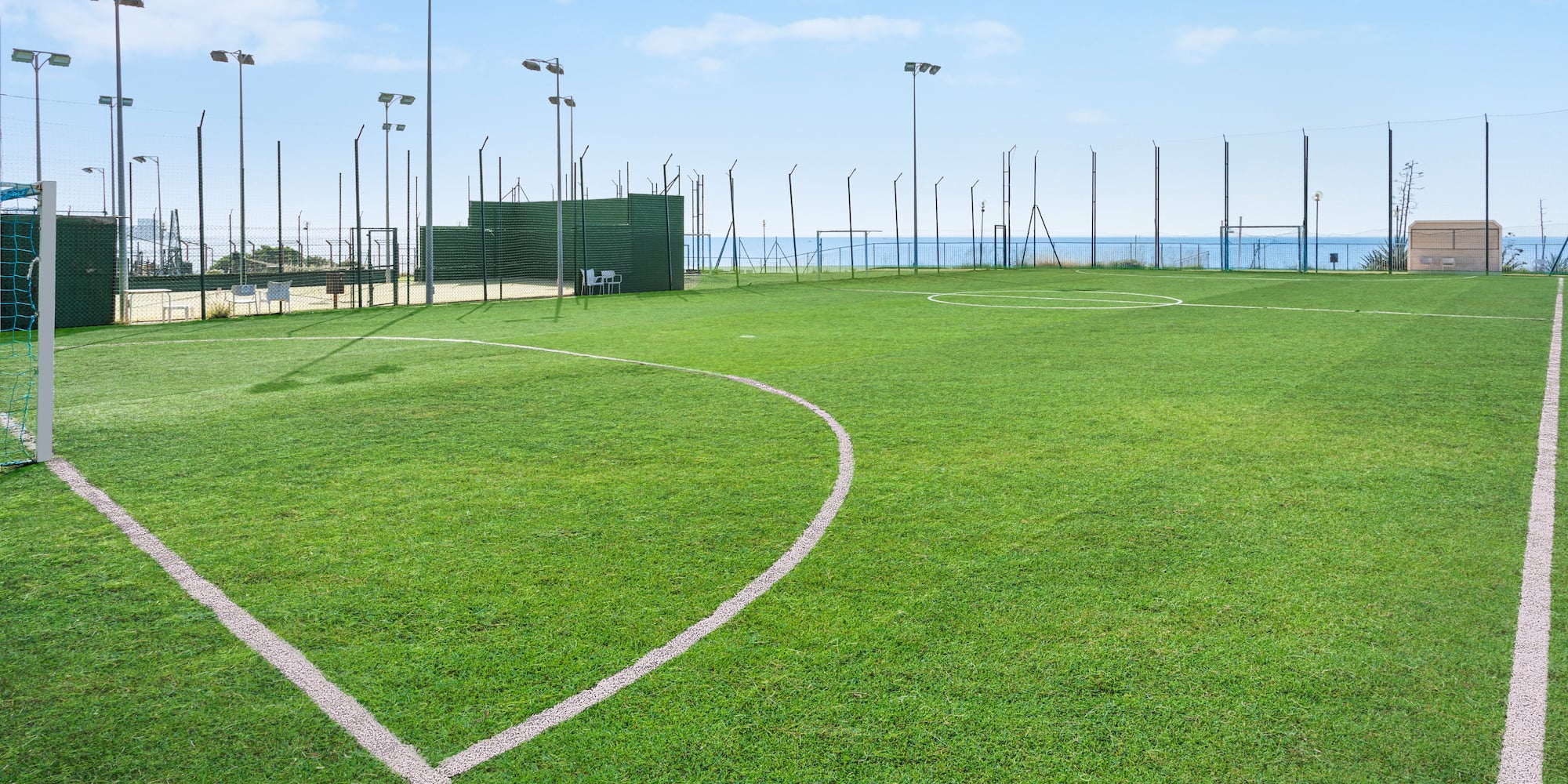 Green artificial turf soccer field with white lines, floodlights, and sea view.