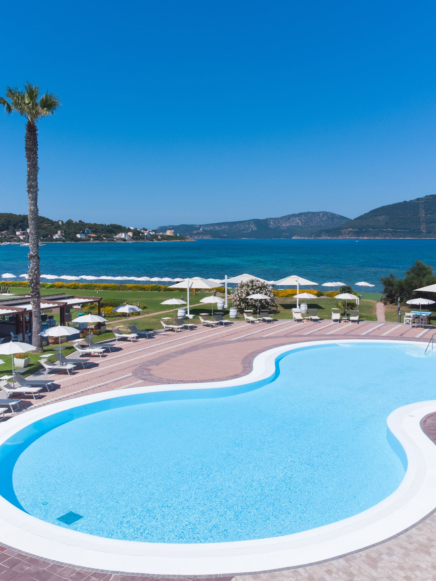Resort swimming pool, blue sea, mountains, palm trees, lounge chairs.