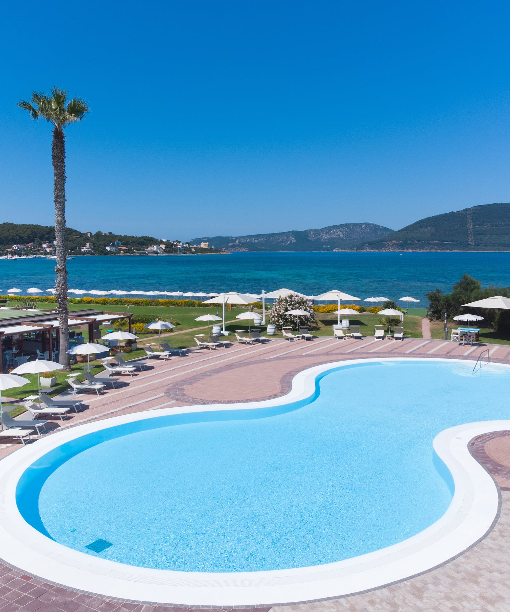 Resort swimming pool, blue sea, mountains, palm trees, lounge chairs.