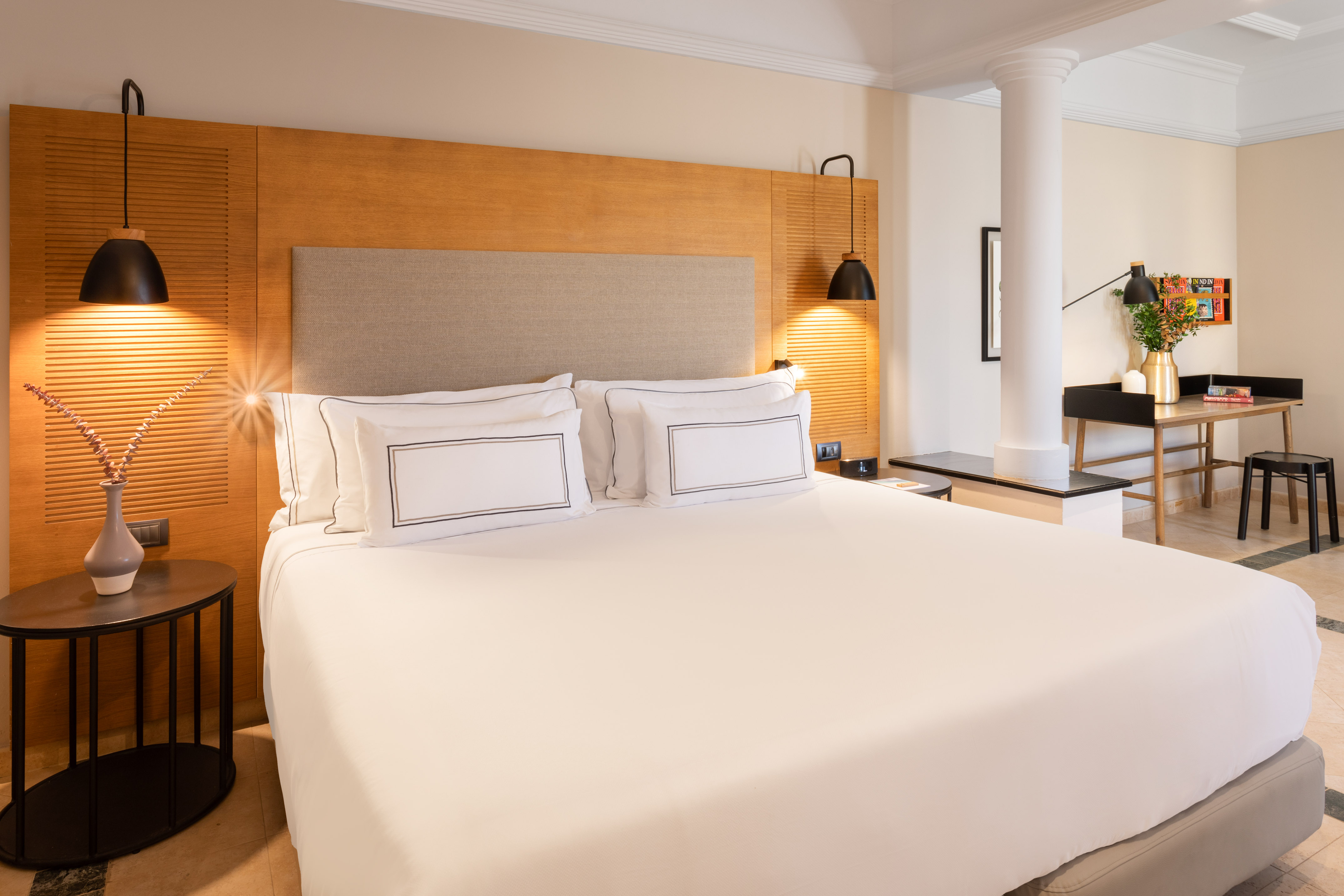 Modern hotel room with large white bed, wood headboard, accent lights, and desk.