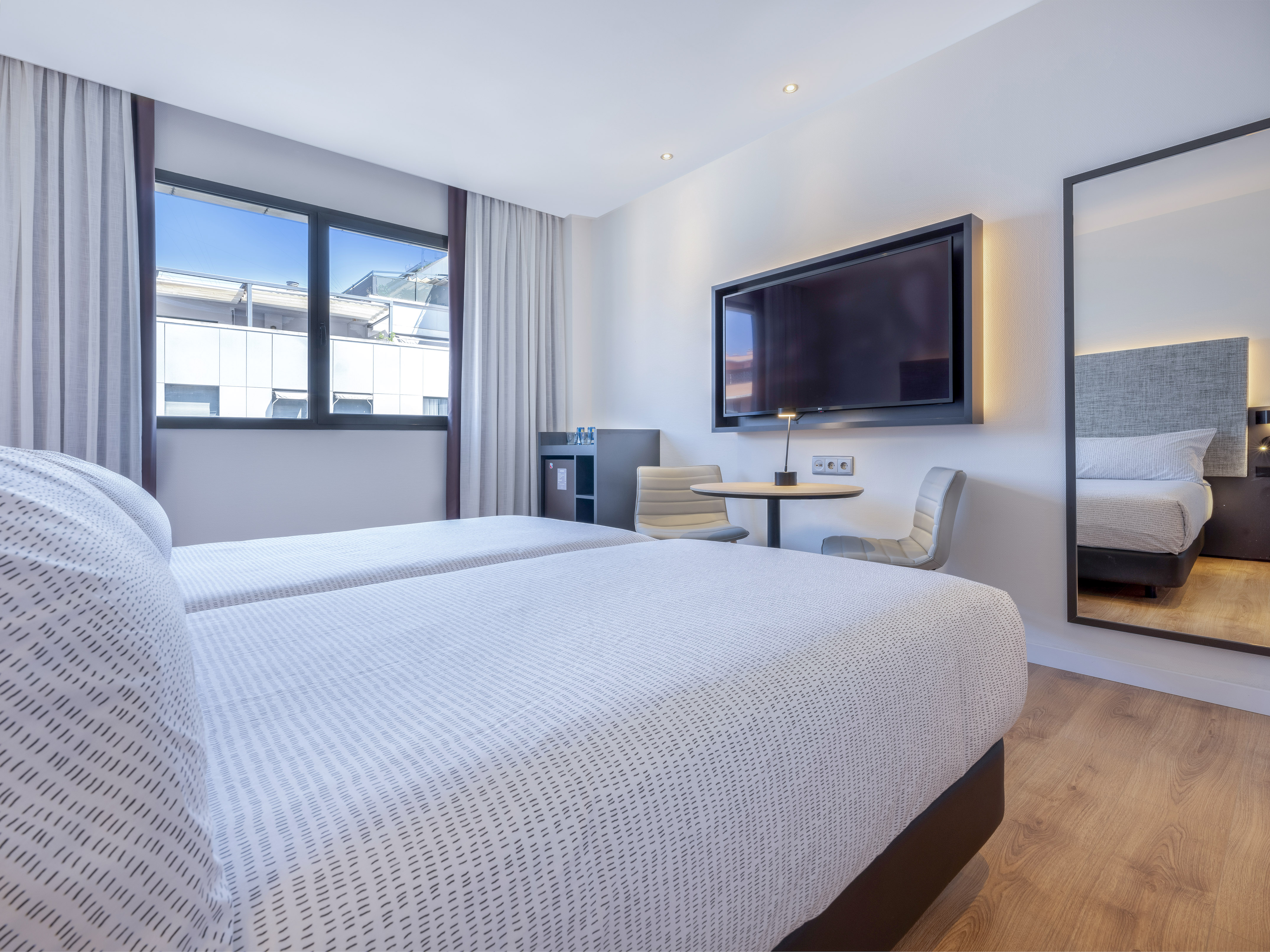 Modern hotel room with twin beds, wall-mounted TV, window, and wood flooring.