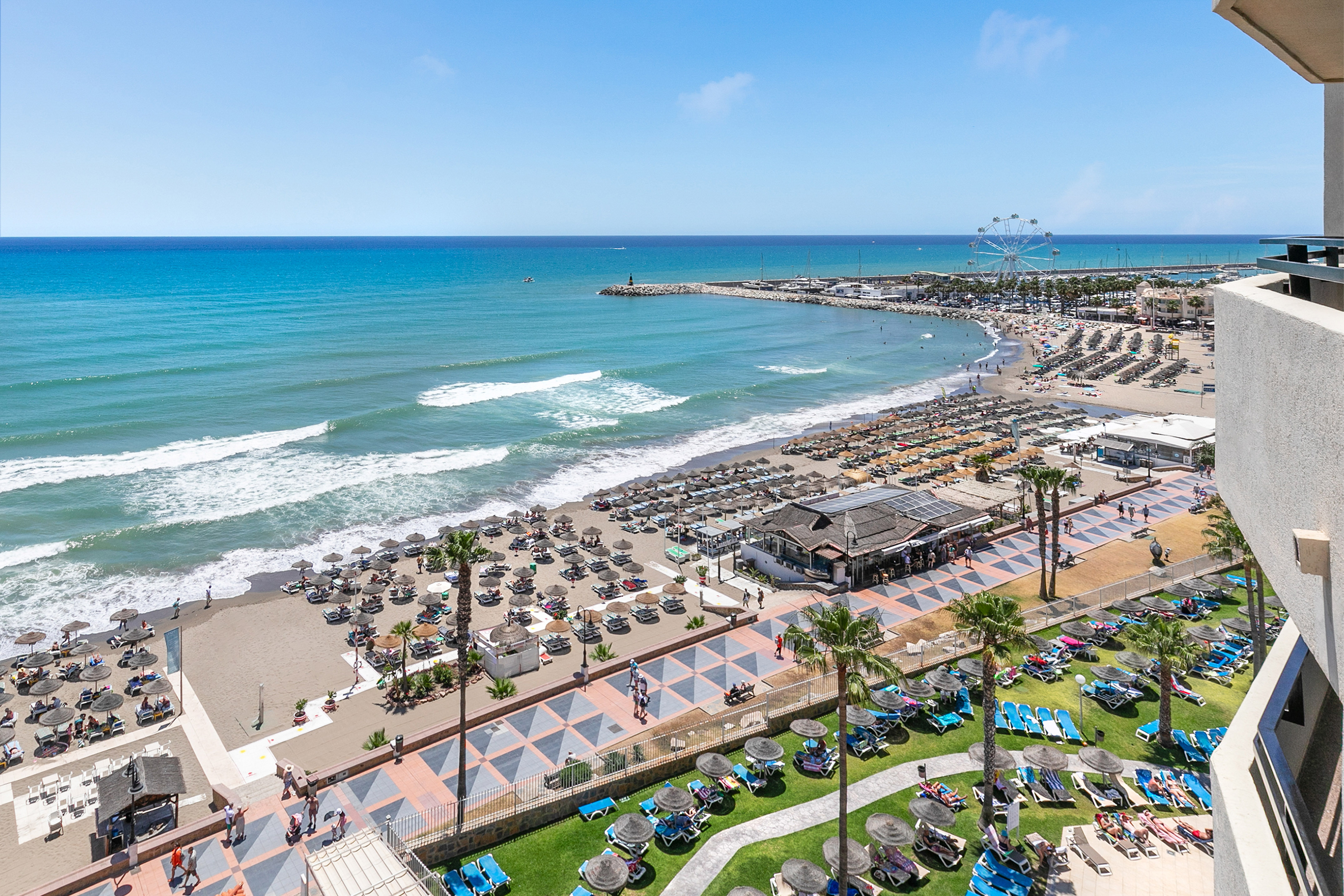 Beach resort with ocean, sandy shore, promenade, umbrellas, and Ferris wheel.