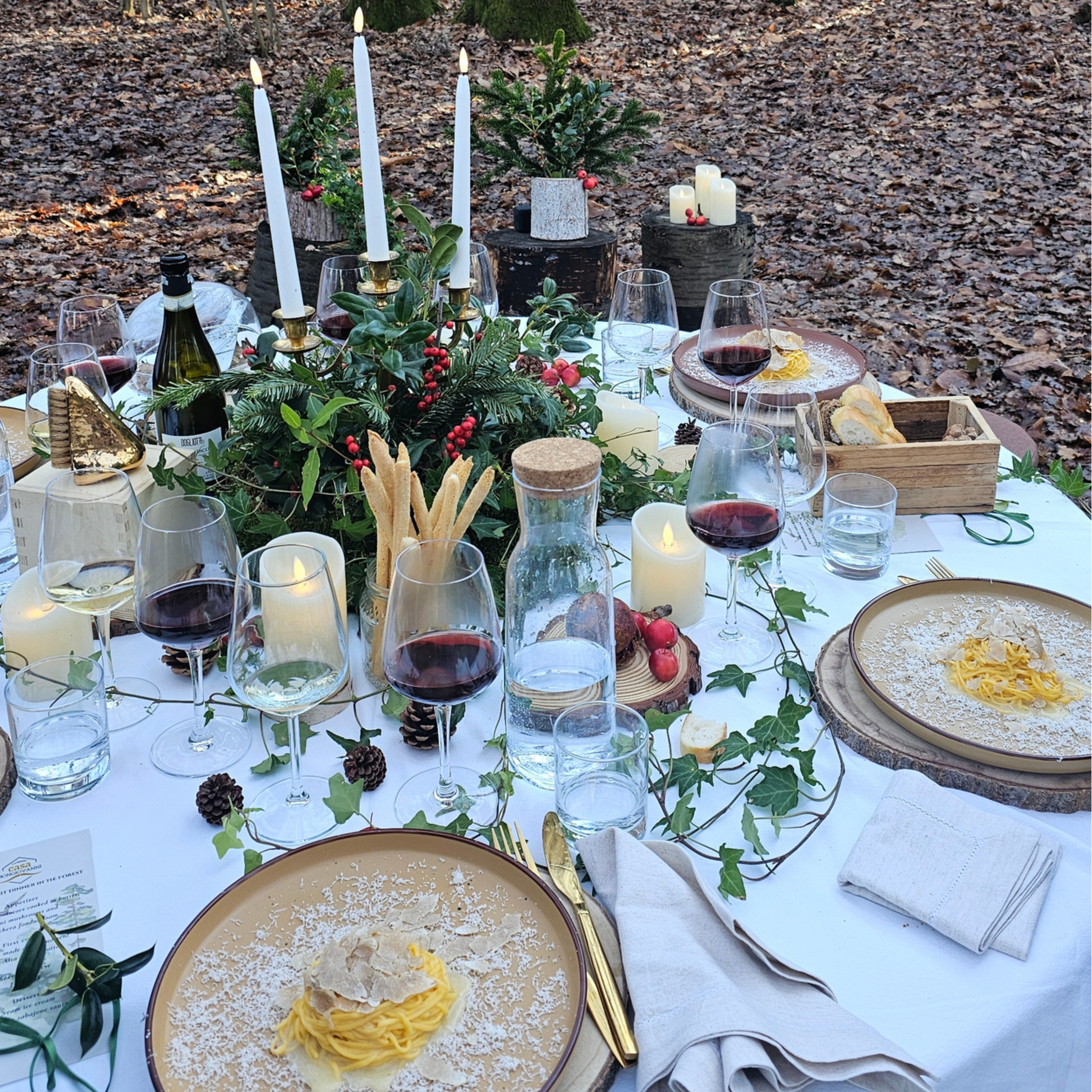 Candlelit forest dinner table with truffle pasta, red wine, and festive decor.