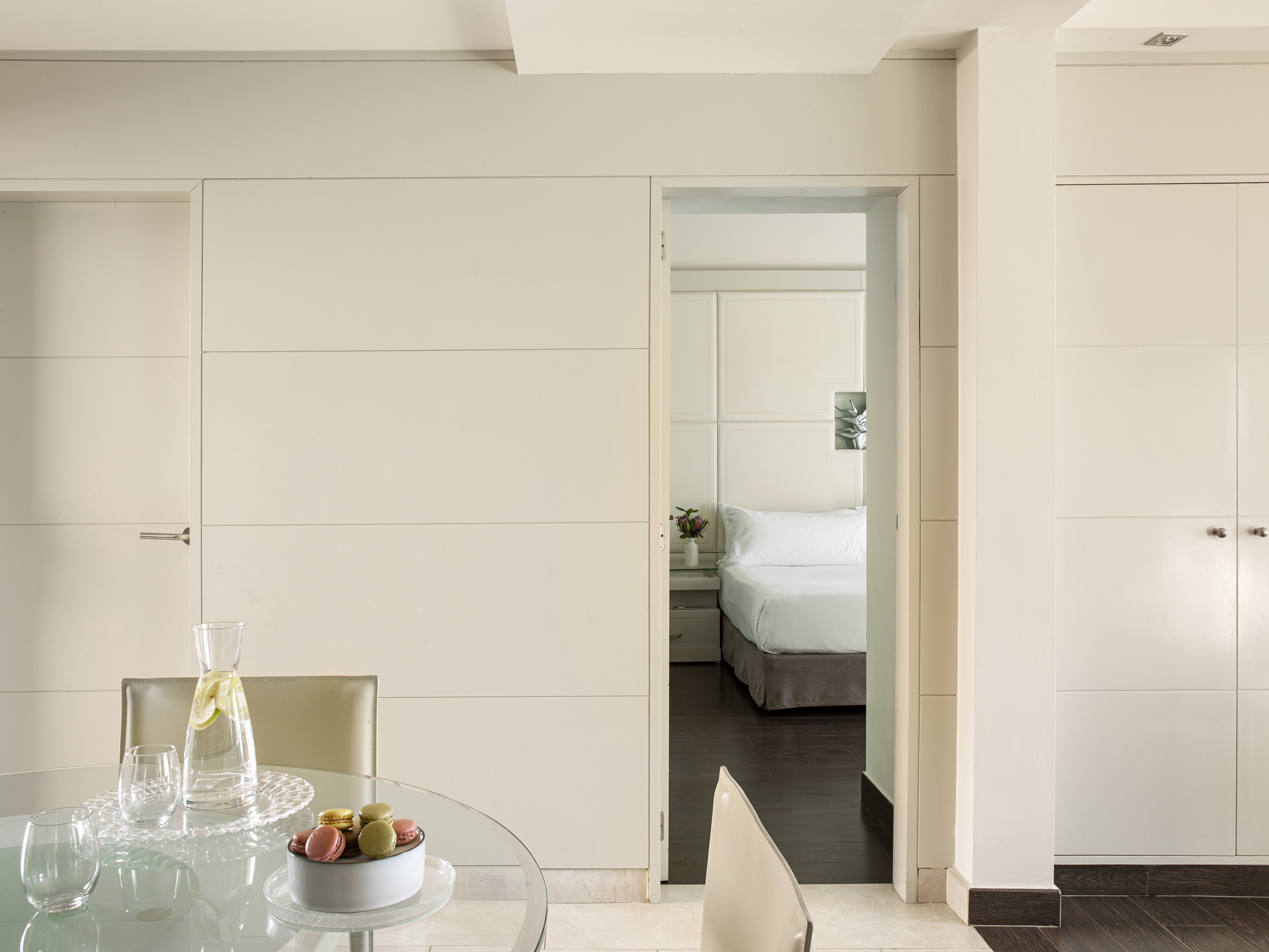 Modern apartment dining area with macarons on glass table, bedroom visible.
