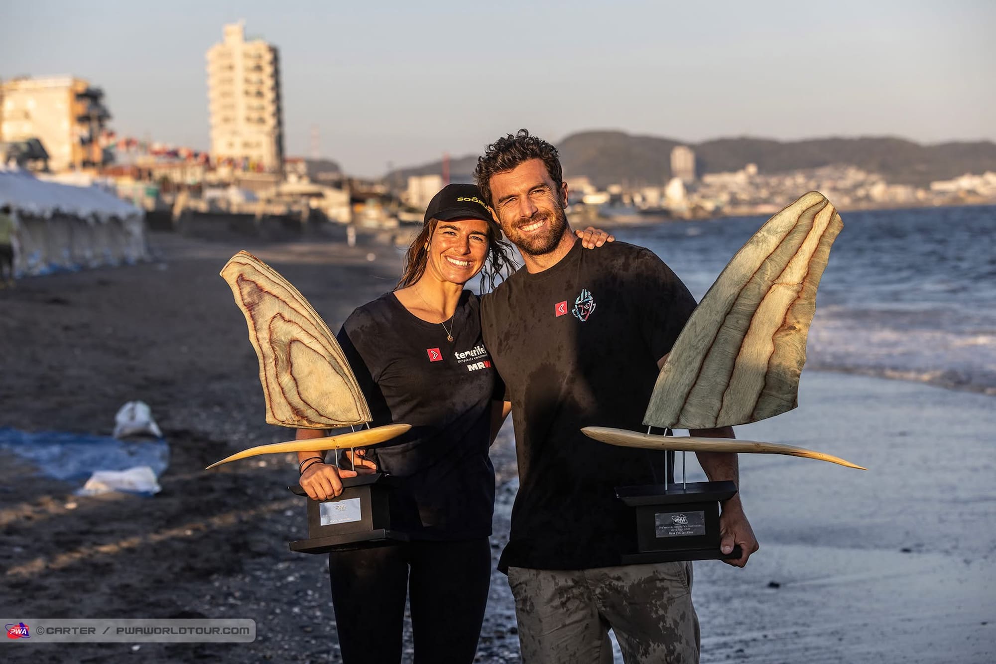 Campeones de windsurf sonriendo con trofeos de aleta en la playa al atardecer.