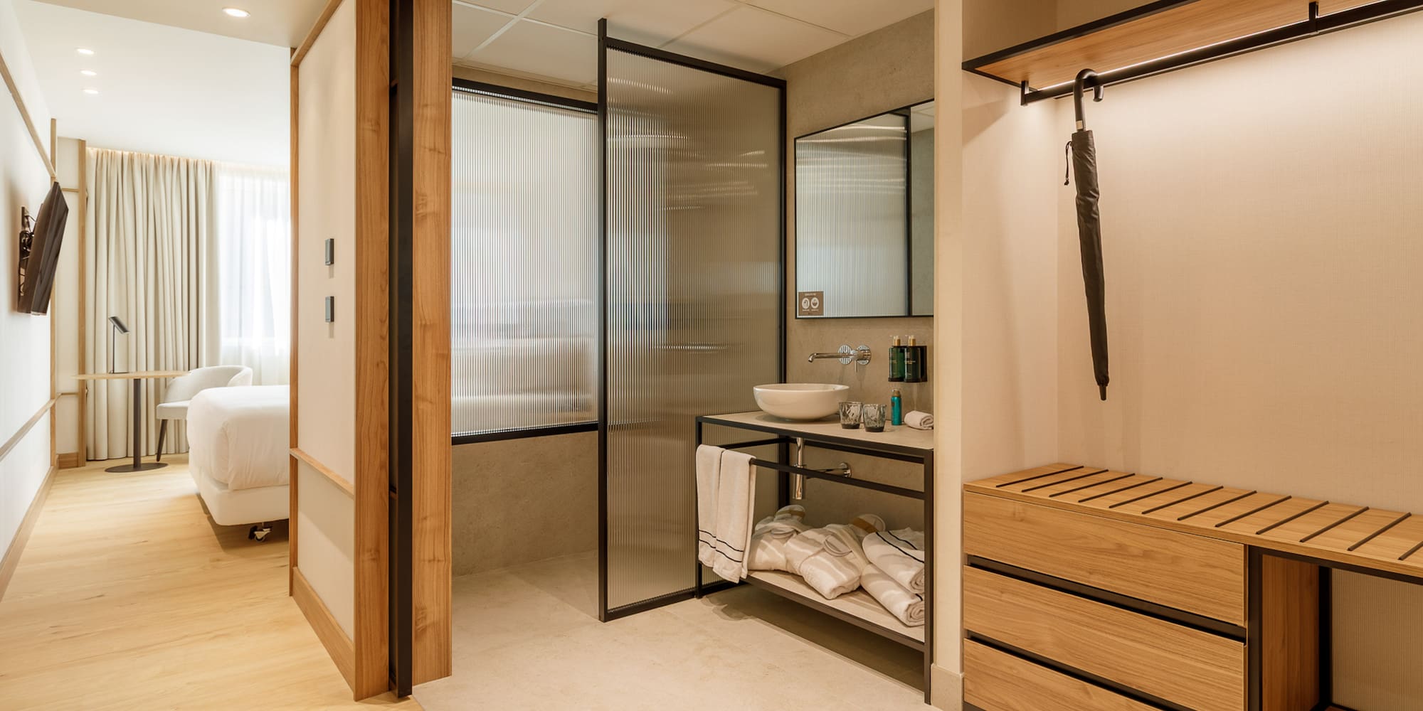 Modern hotel room: open bathroom, fluted glass, wood vanity & storage.