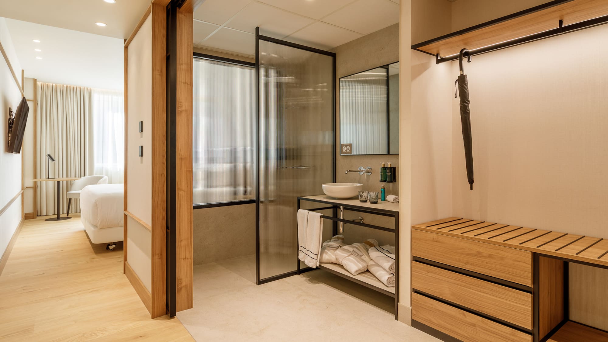 Modern hotel room with open bathroom, fluted glass shower, and wood storage.