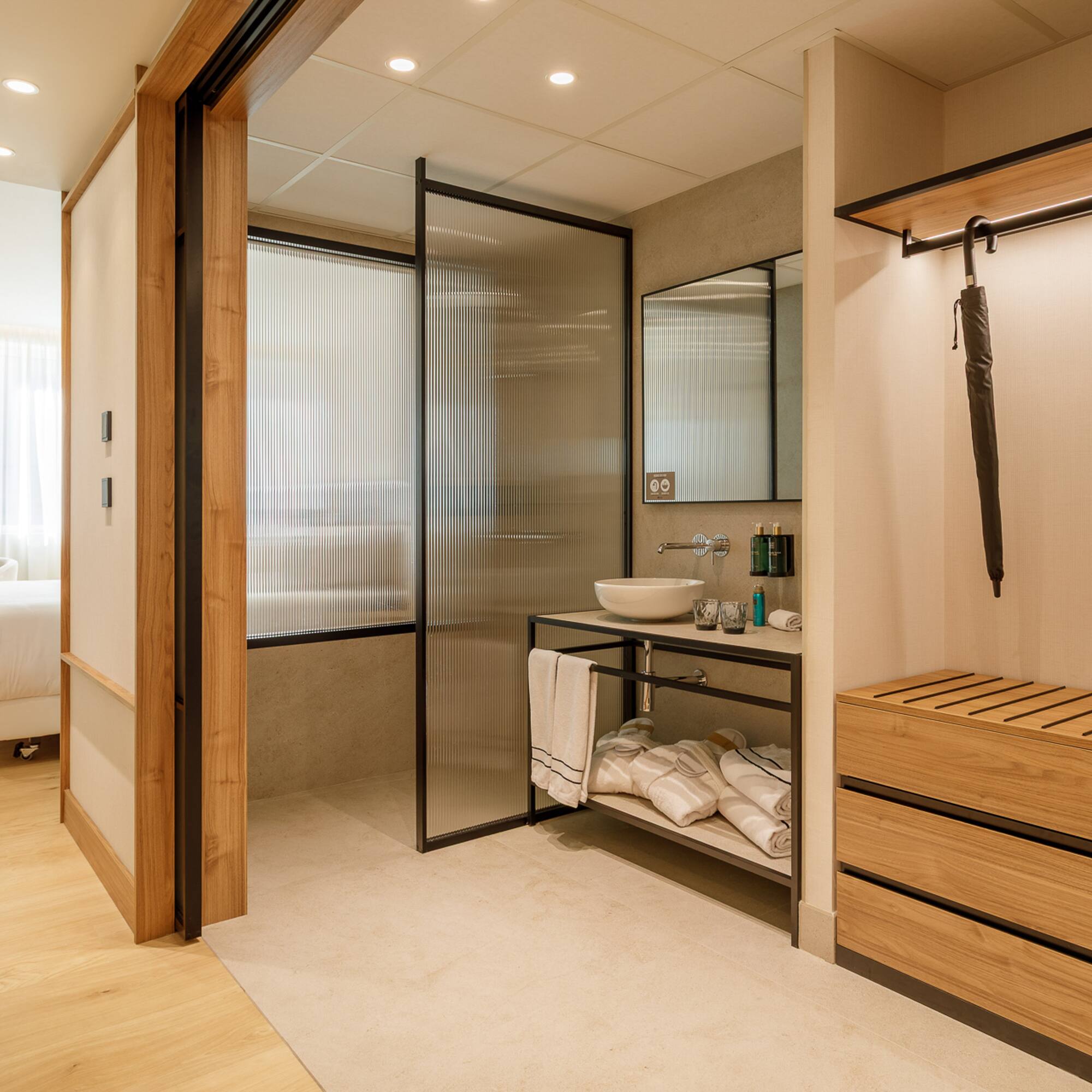 Modern hotel room: open bathroom, fluted glass, wood vanity & storage.