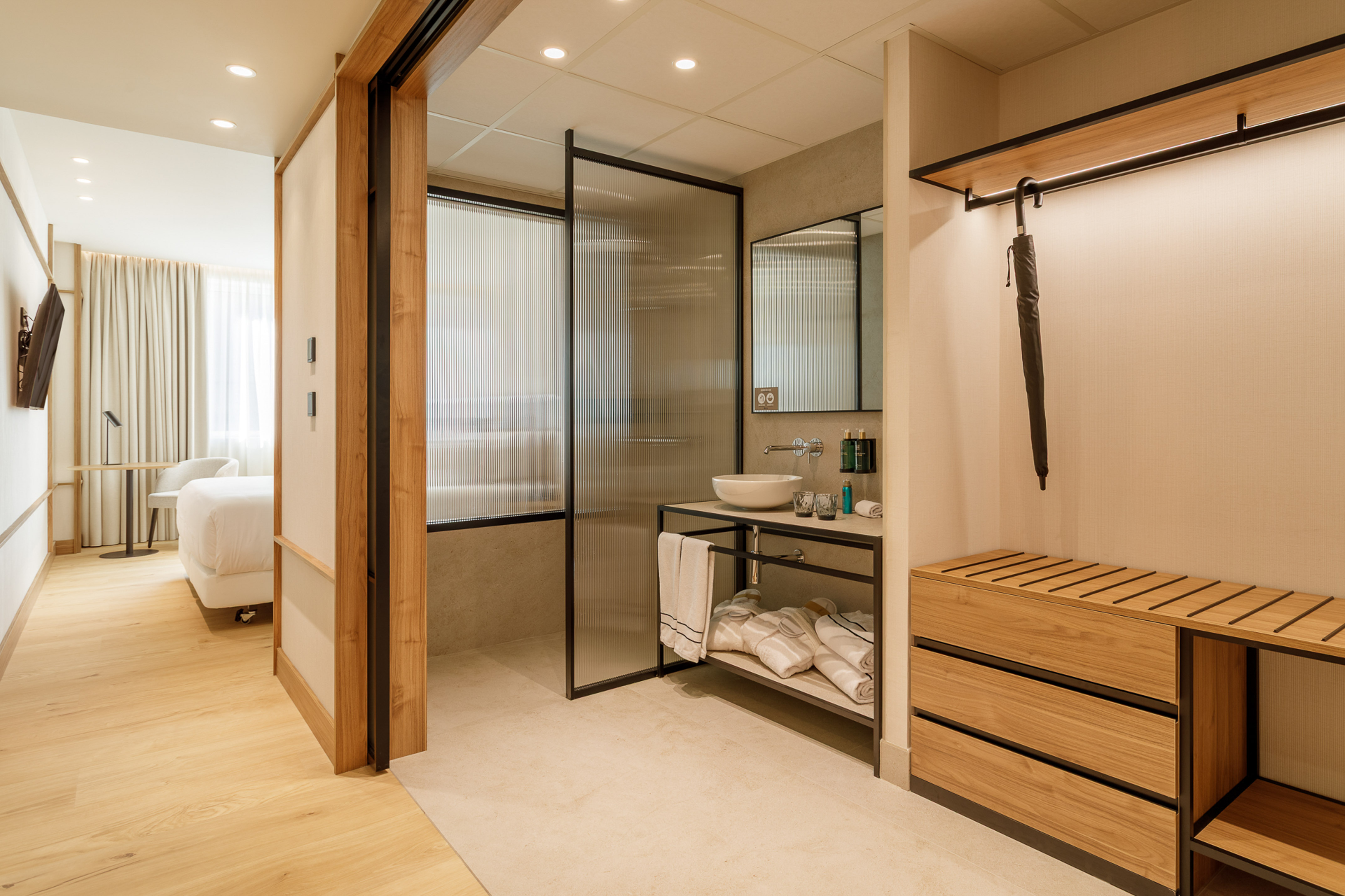 Modern hotel room with open bathroom, fluted glass shower, and wood storage.