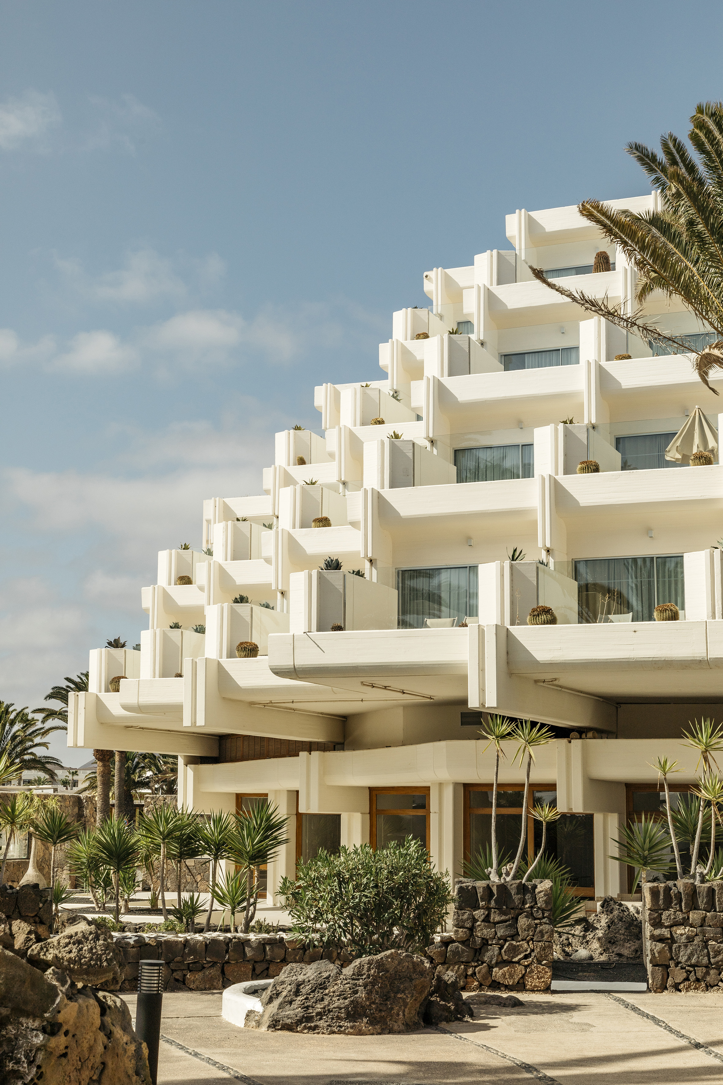 Modern white tiered resort with balconies, cacti, and palm trees.