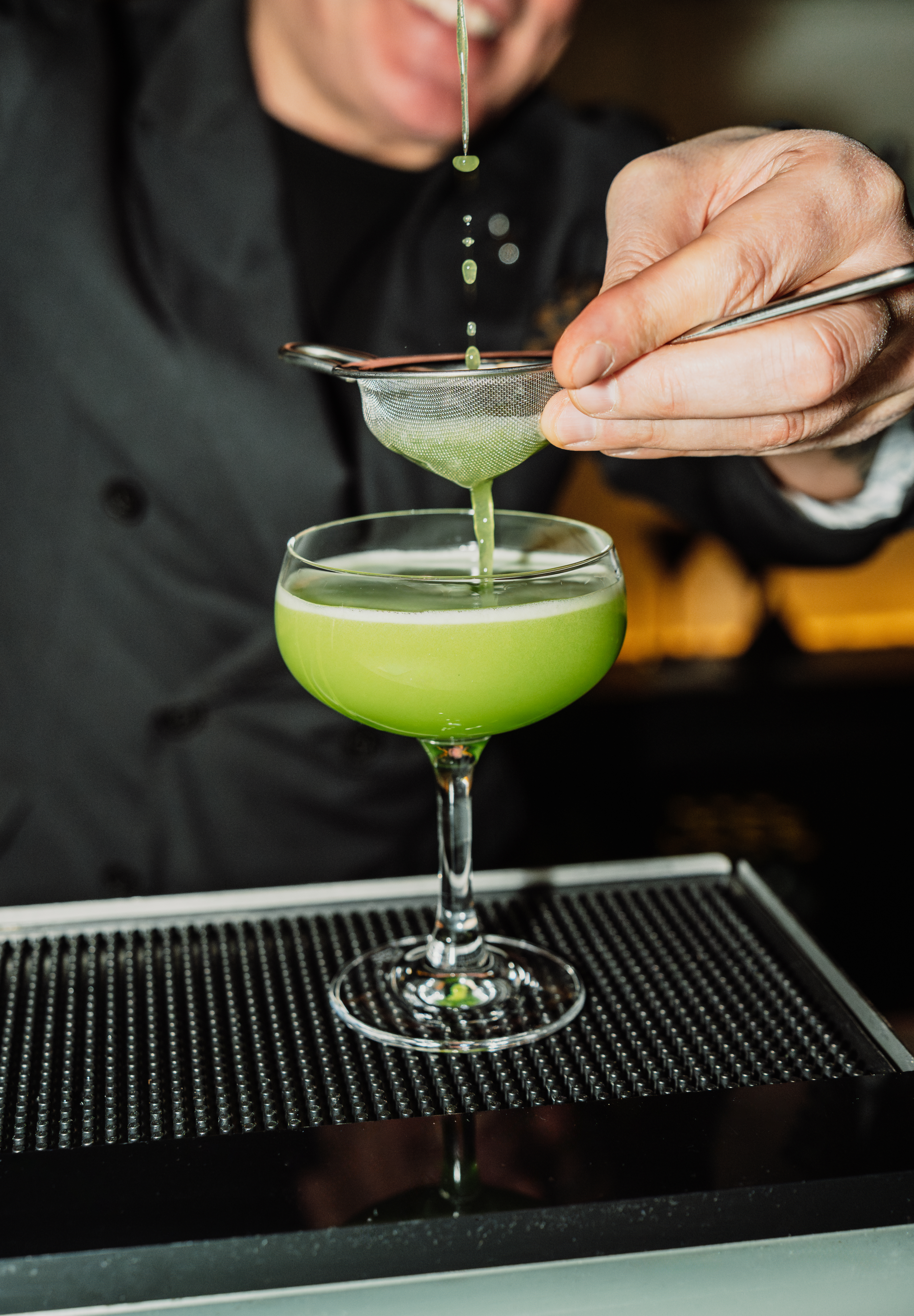 Bartender straining a green cocktail into a coupe glass.