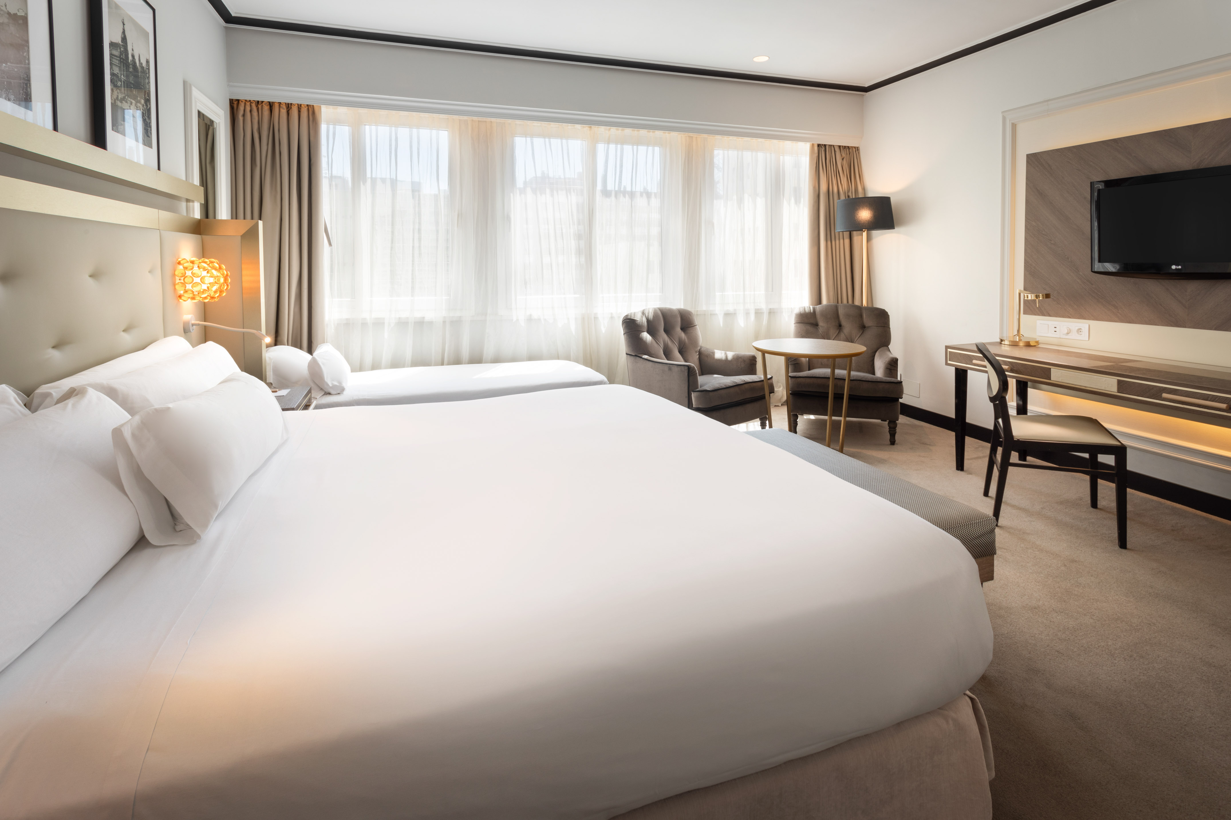 Elegant hotel room with twin beds, bright window, desk, TV, and sitting area.