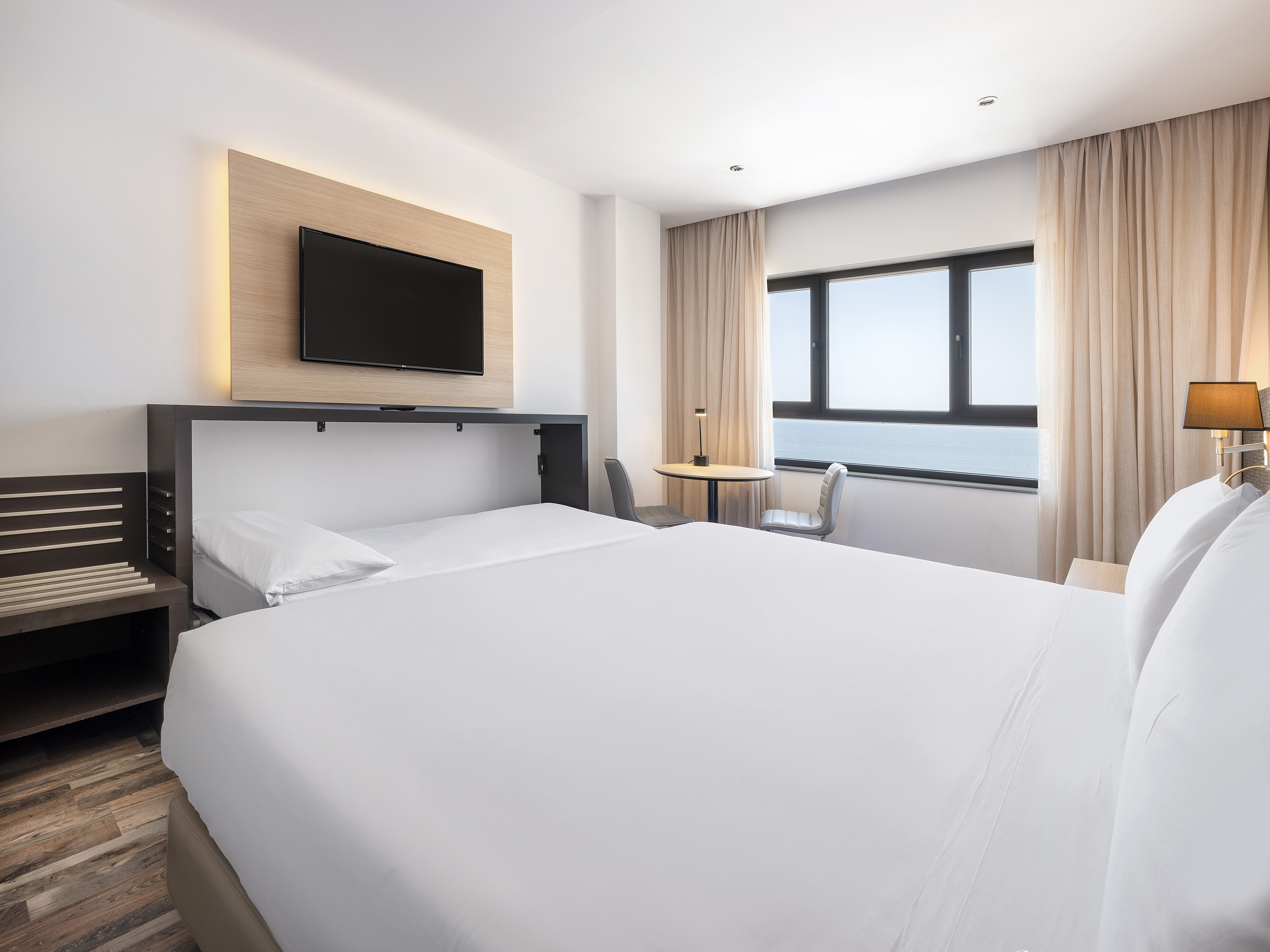 Bright hotel room with white bed, wall-mounted TV, and ocean view.