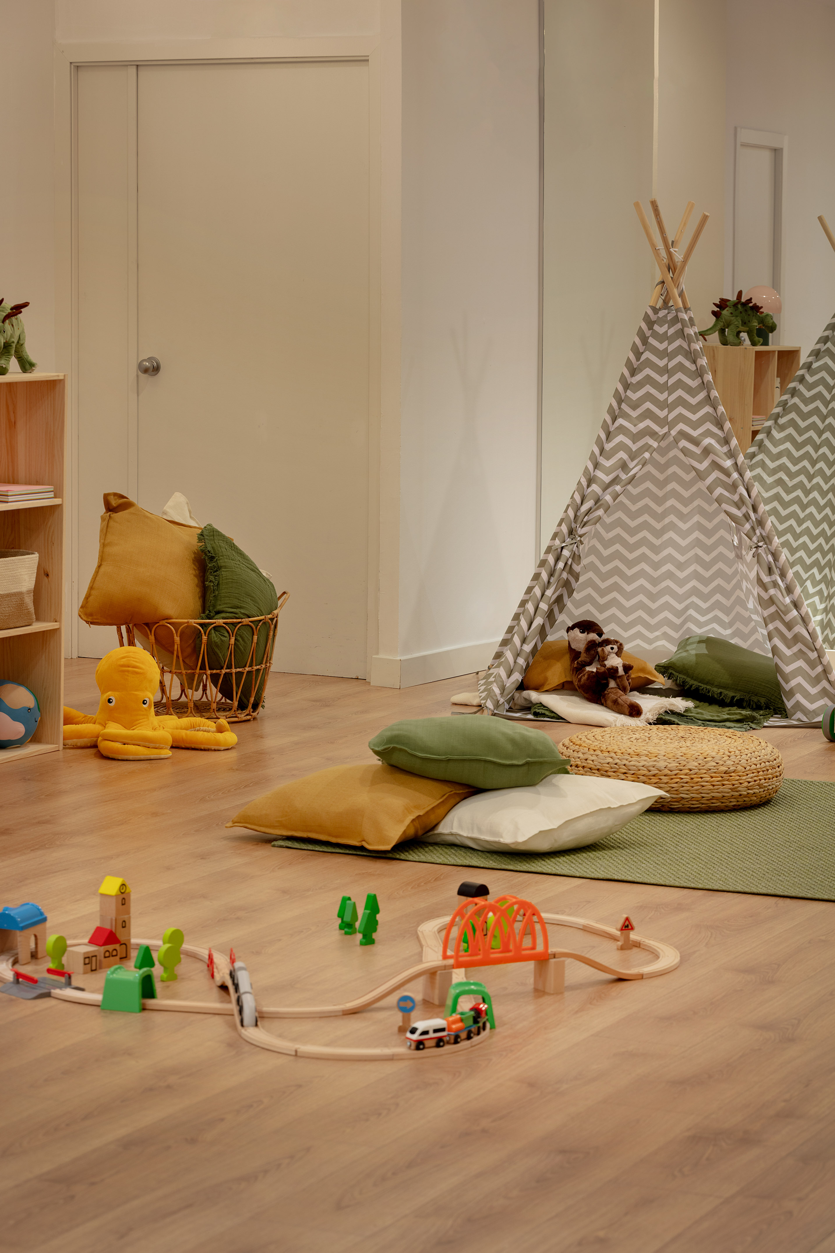 Playroom with grey teepee, wooden train set, plush toys, and floor pillows.