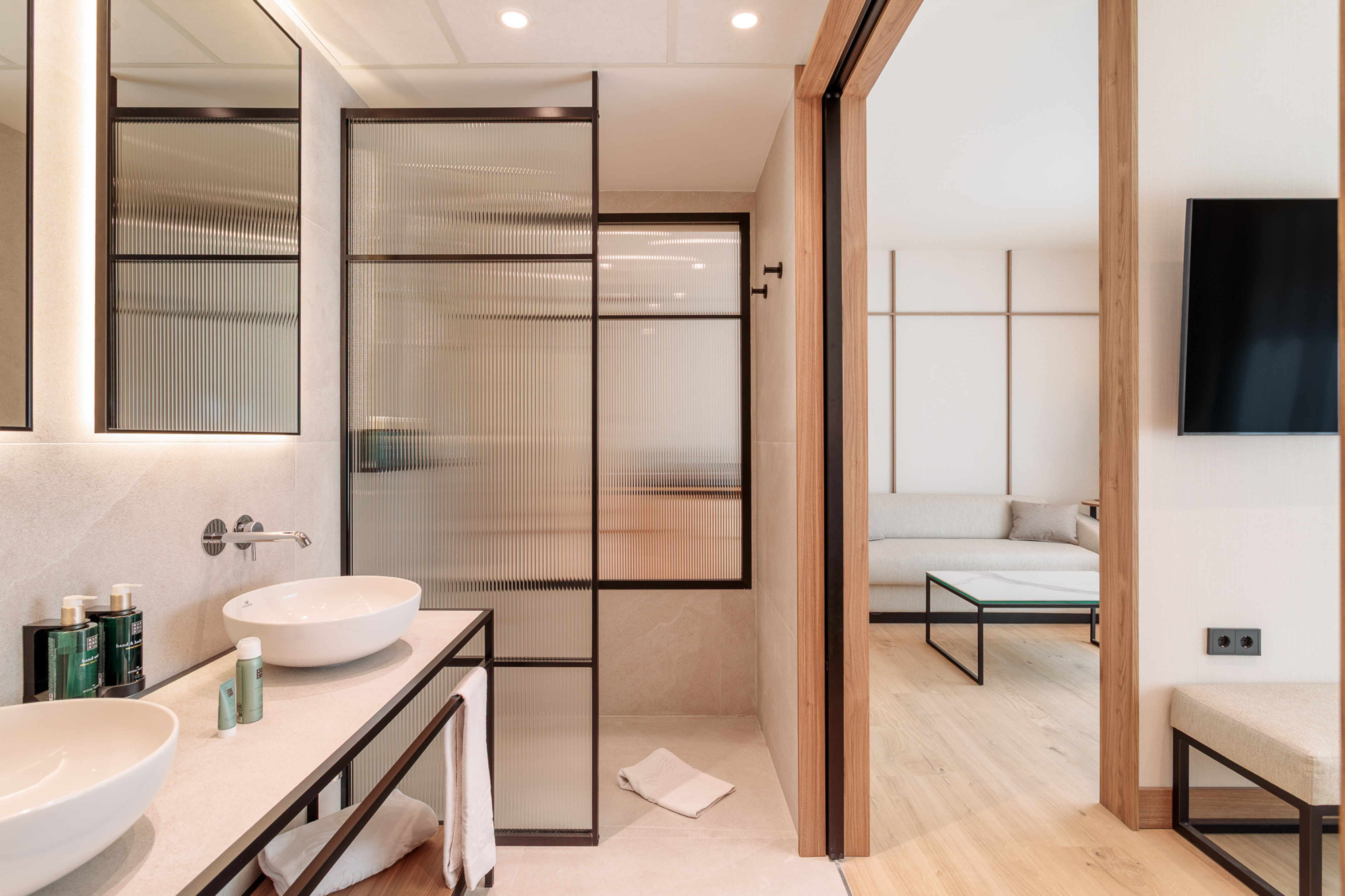 Modern bathroom with double sinks, fluted glass shower, adjoining living room.