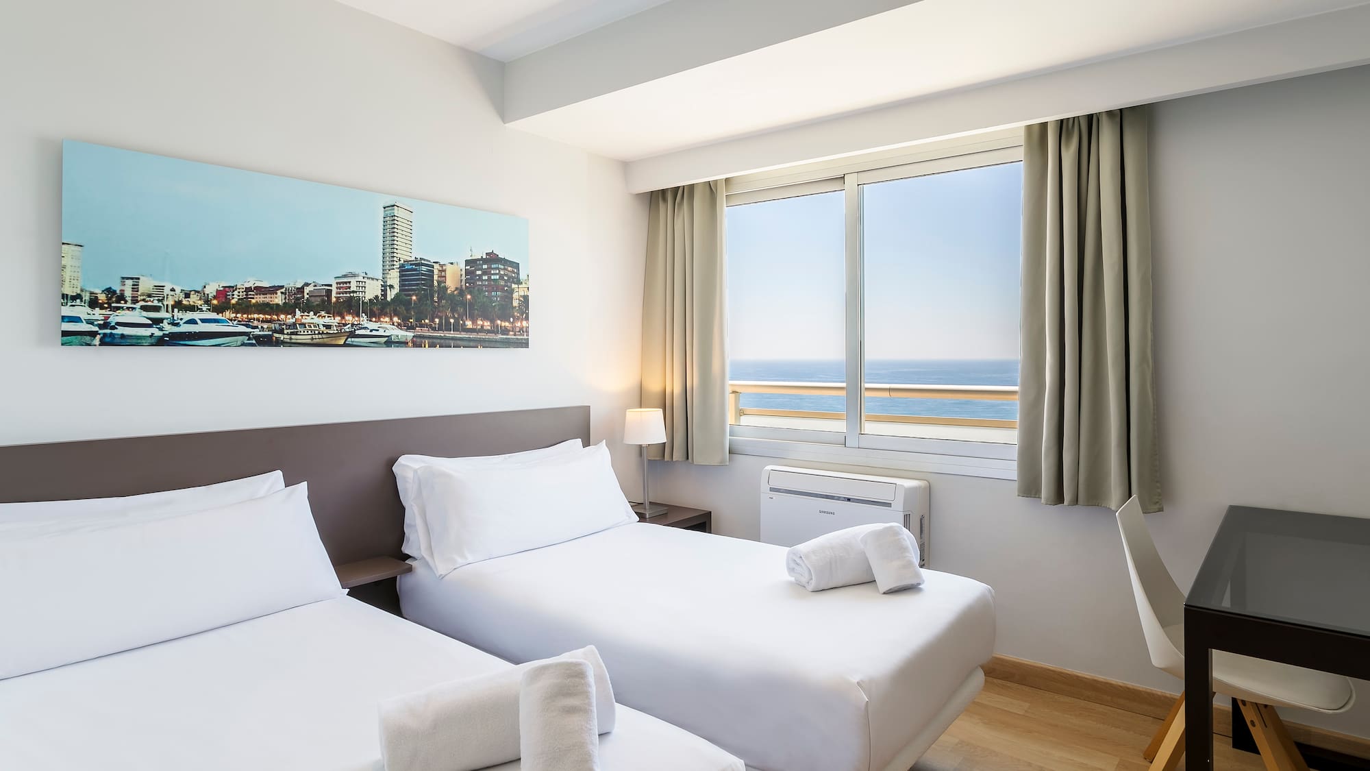 Modern hotel room with twin beds, ocean view, city skyline art, and a work desk.