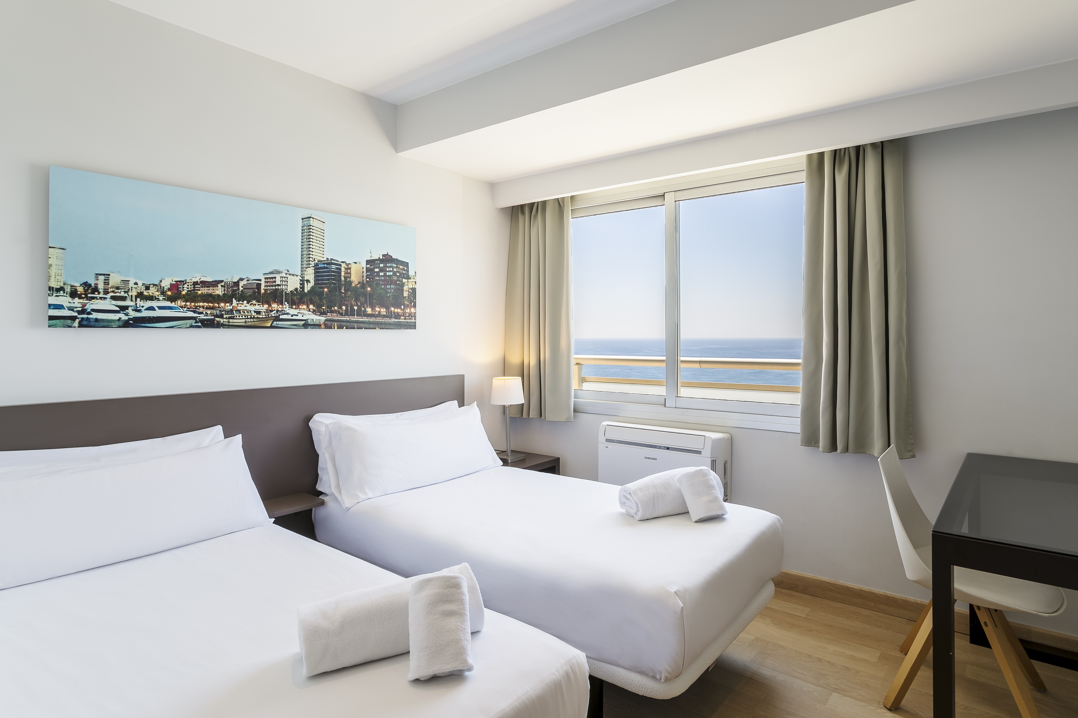 Modern hotel room with twin beds, ocean view, city skyline art, and a work desk.