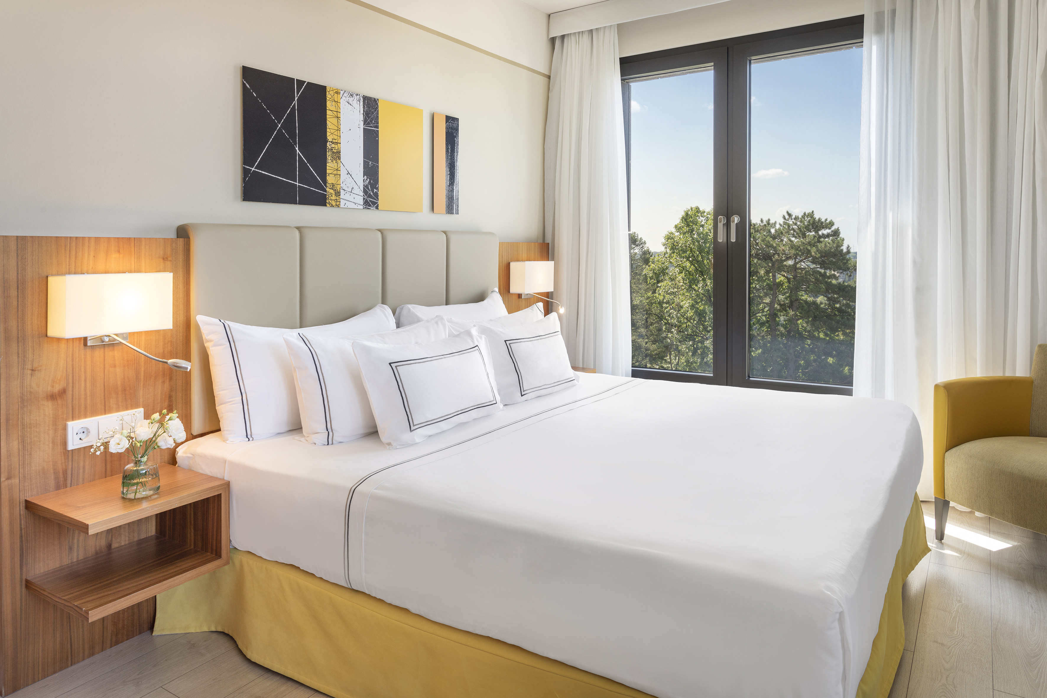 Modern hotel room with white bed, wood accents, yellow details, and window view.