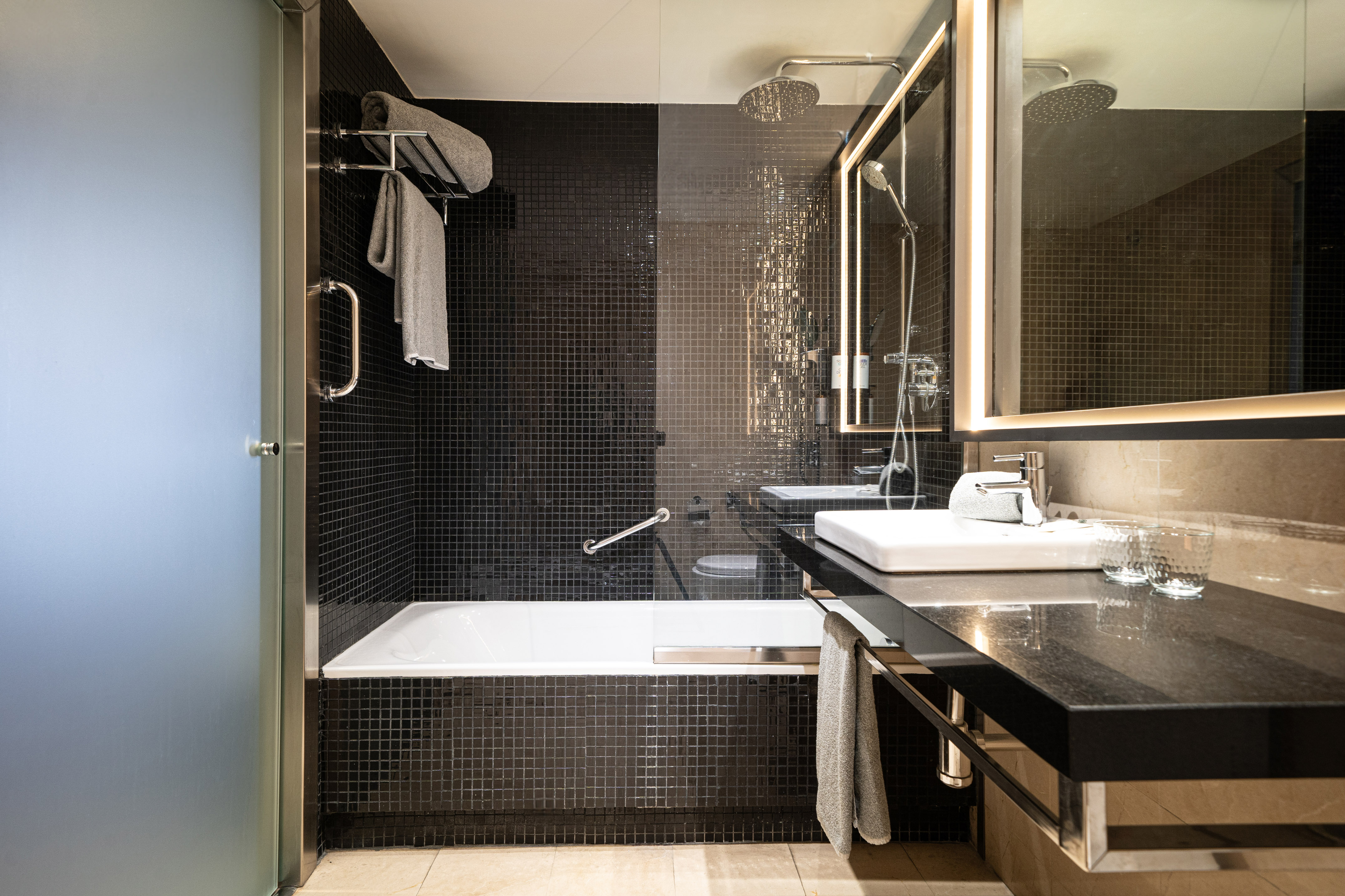 Modern bathroom with black tiles, shower/tub, vanity, and illuminated mirror.