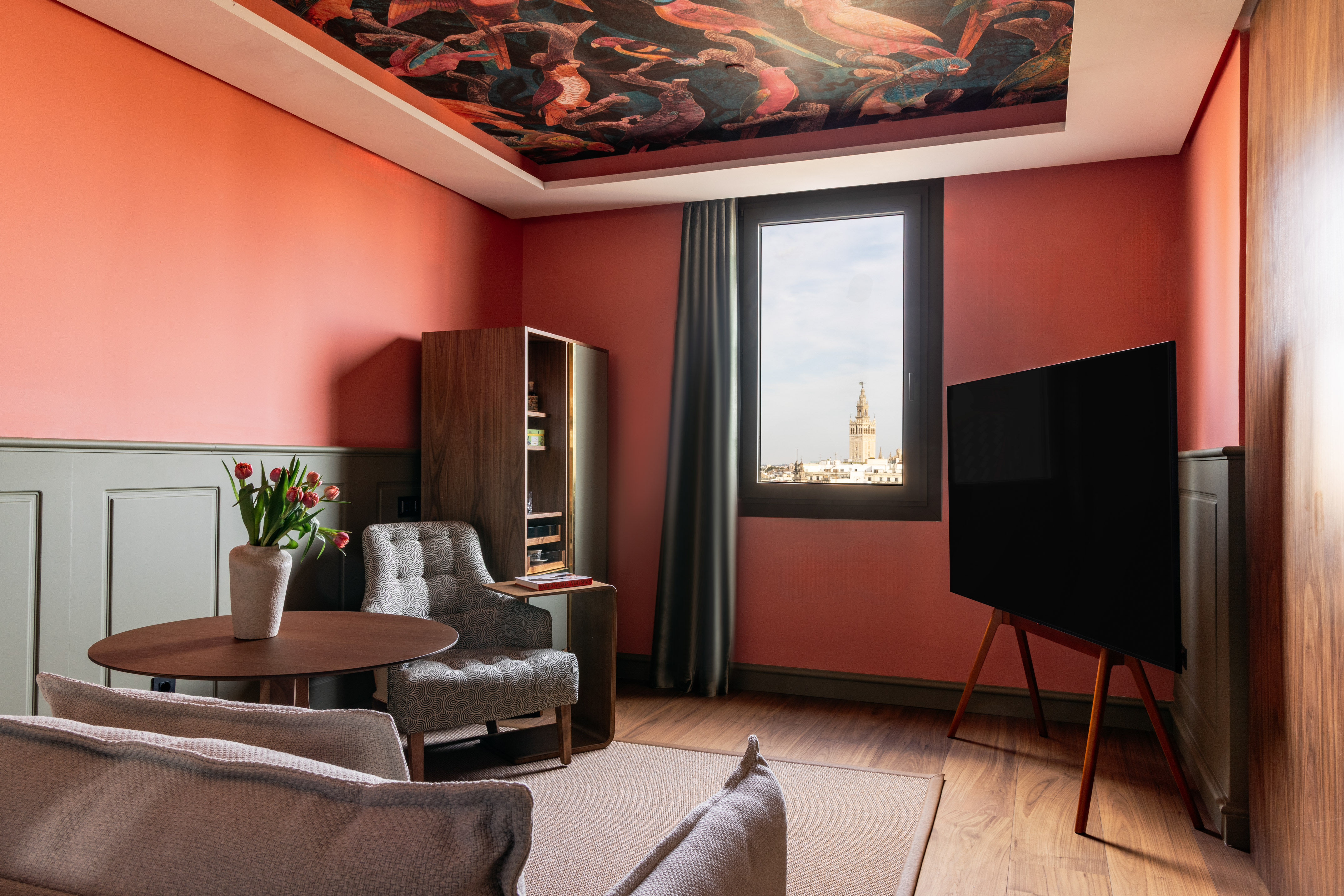 Modern hotel room with coral walls, bird ceiling mural, and Seville Giralda view.