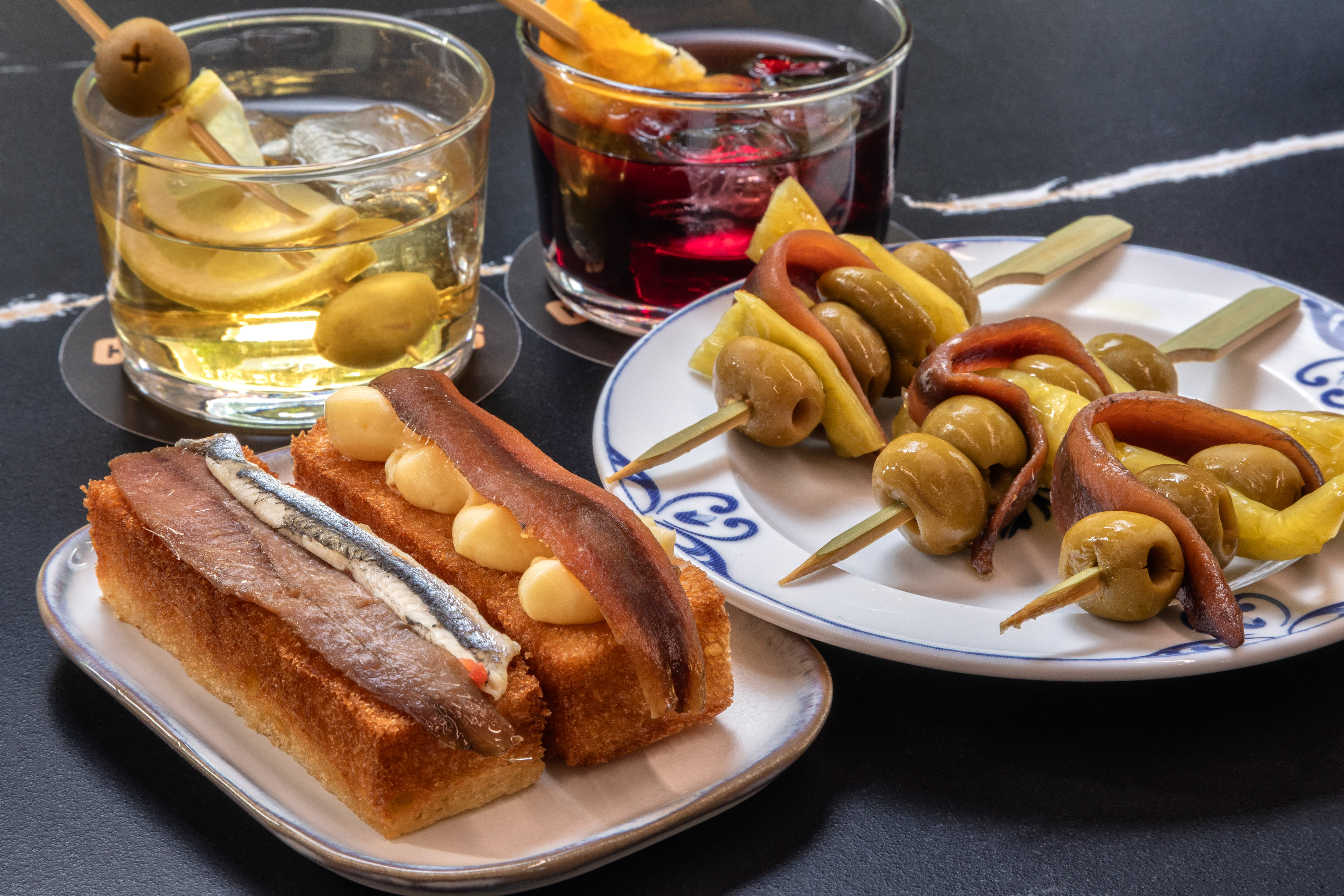 Spanish tapas spread: anchovy pintxos, Gilda skewers, and cocktails on a dark table.