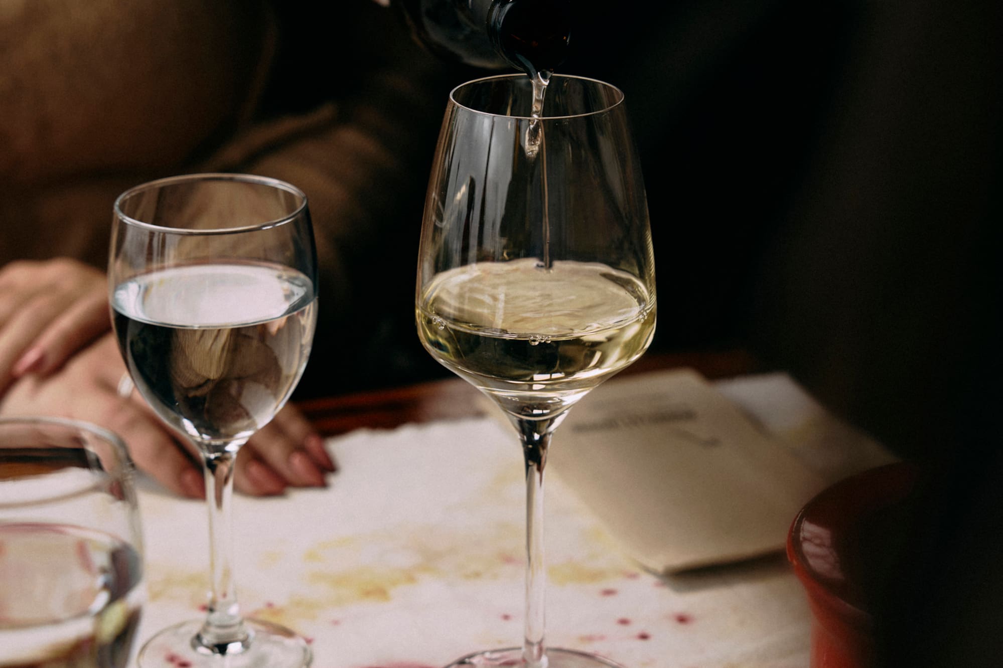 Mano sirviendo vino blanco en copa, mesa con mantel manchado de vino.