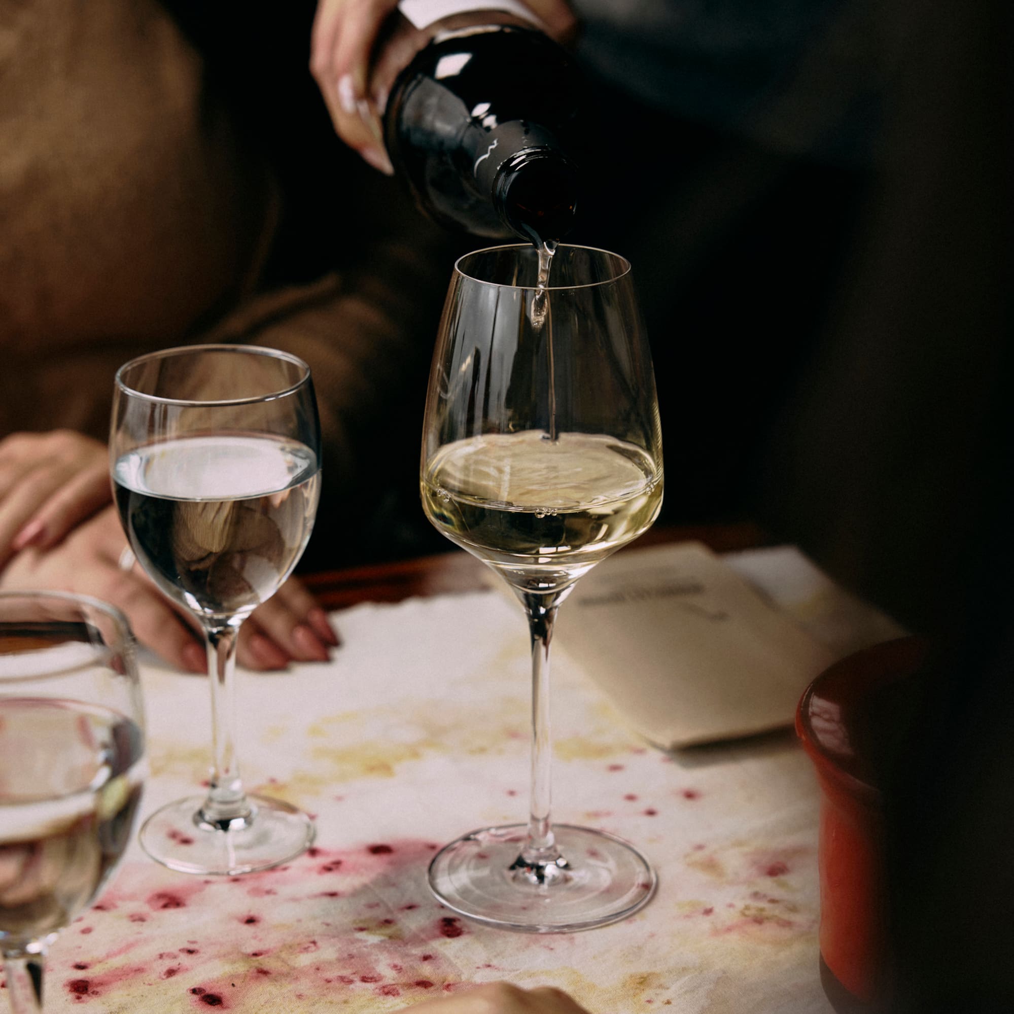 Hand pouring white wine from bottle into glass on a stained dinner table.