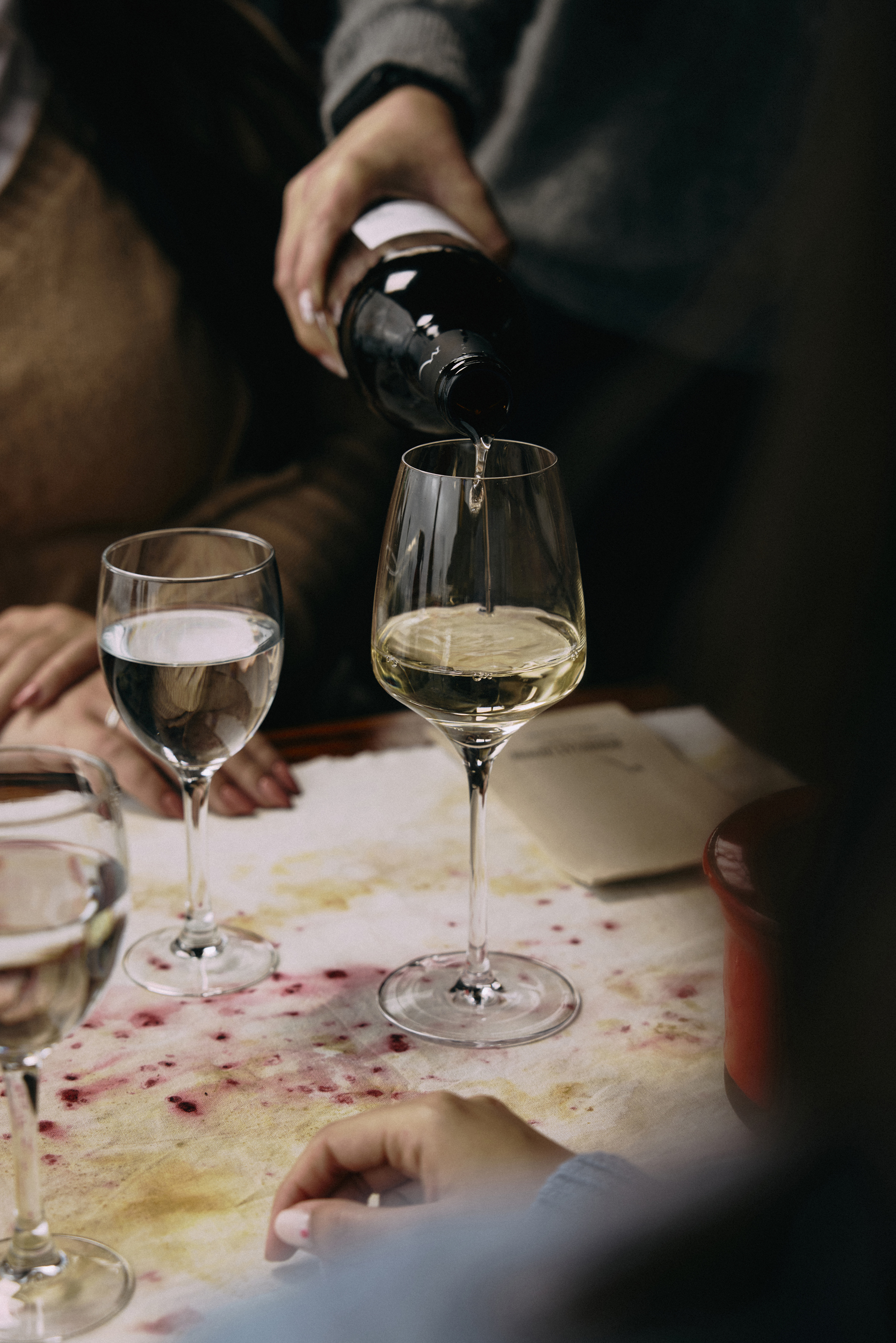 Hand pouring white wine from bottle into glass on a stained dinner table.