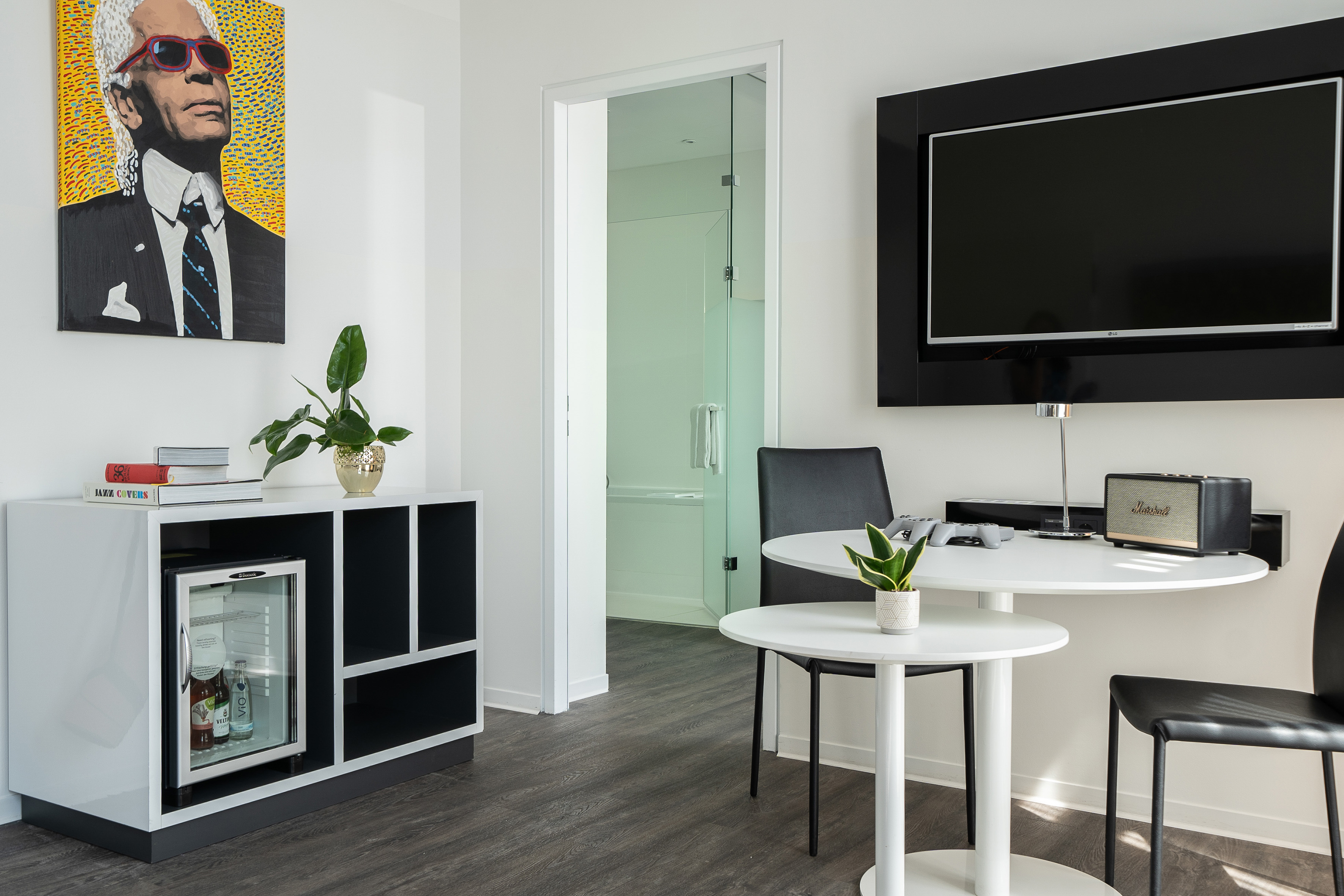 Modern hotel room with Karl Lagerfeld art, TV, mini-fridge, and dining area.