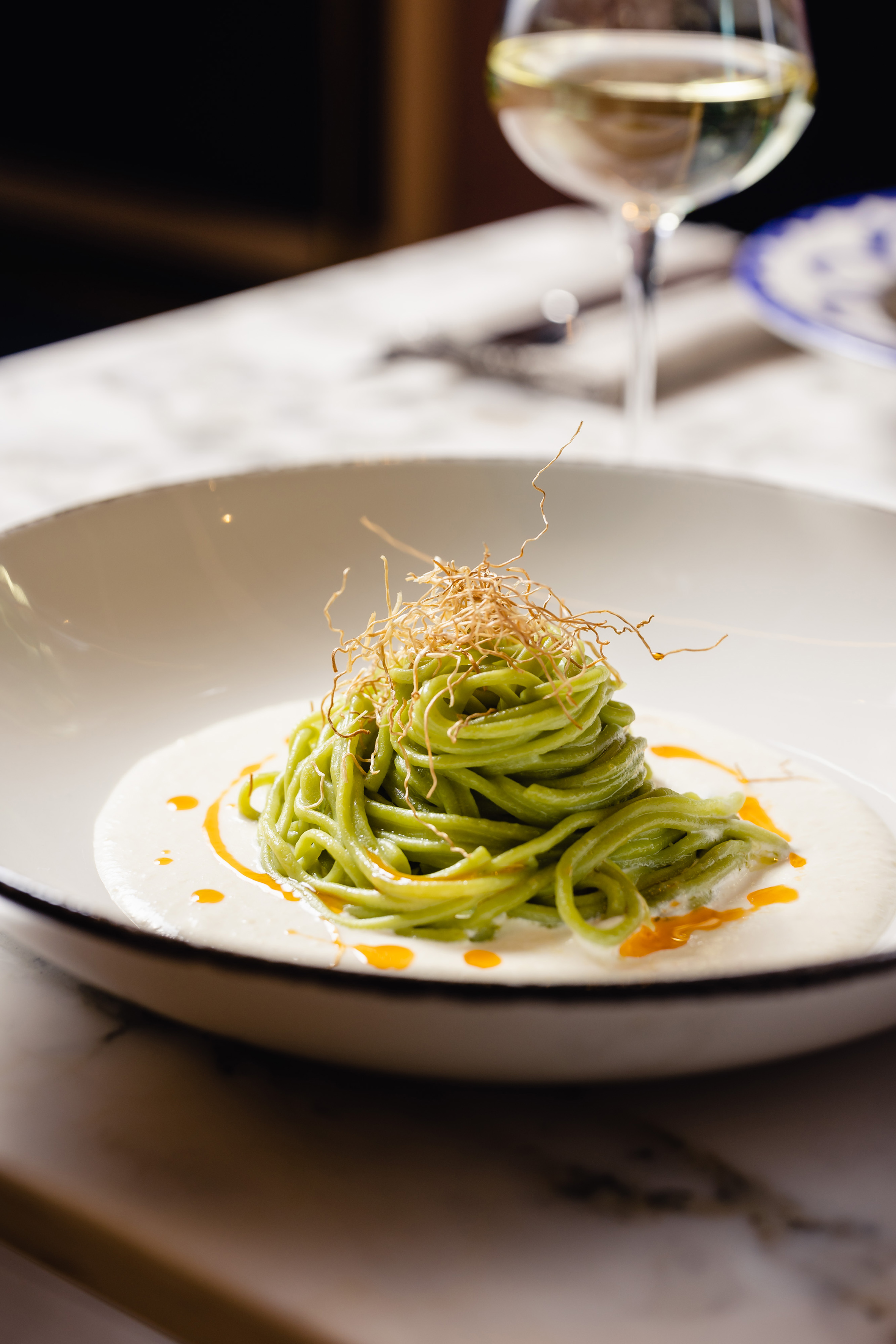 Gourmet green pasta, creamy sauce, crispy topping in white bowl, wine glass.