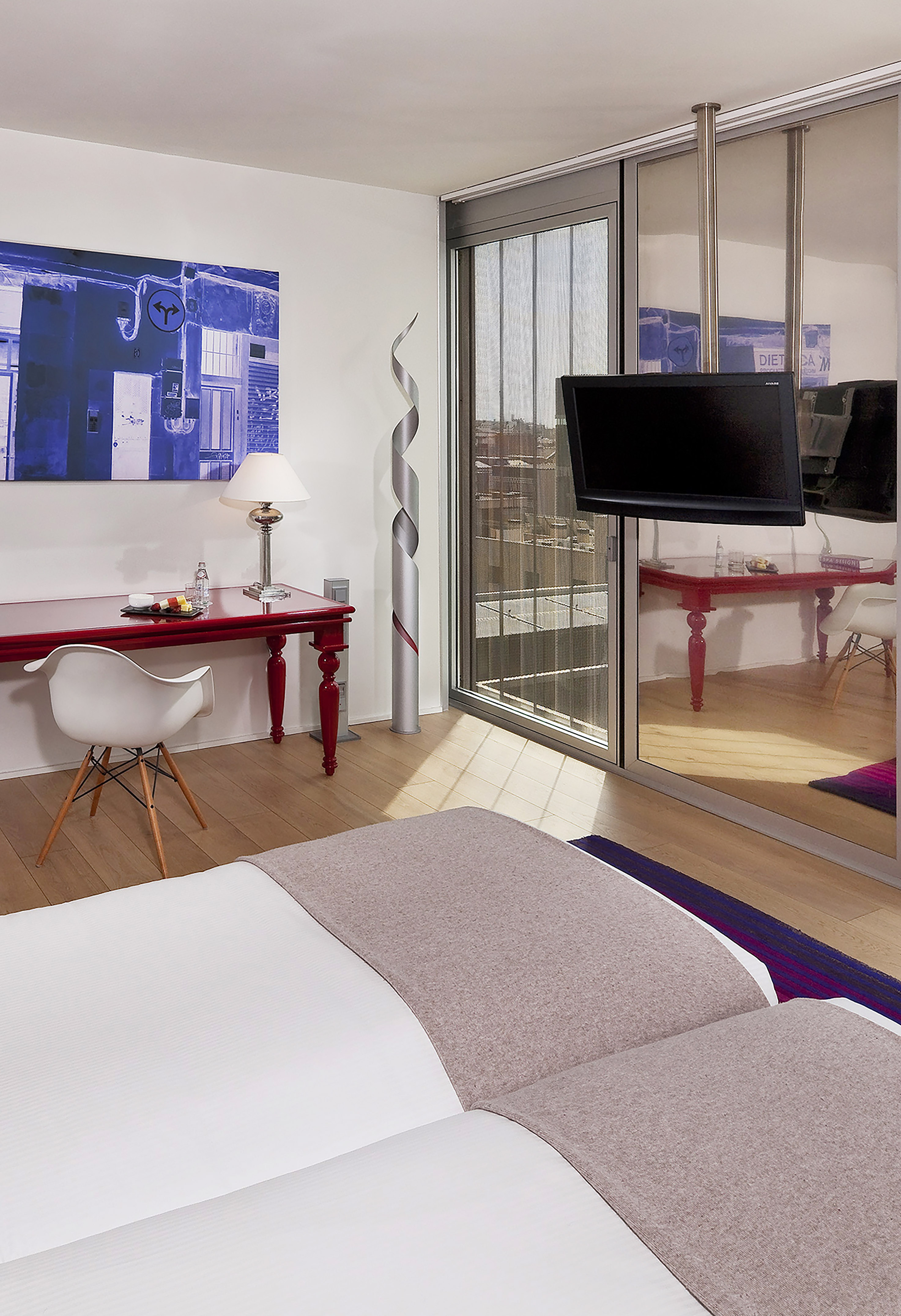 Modern hotel bedroom with white bed, red desk, wall art, and mounted TV.