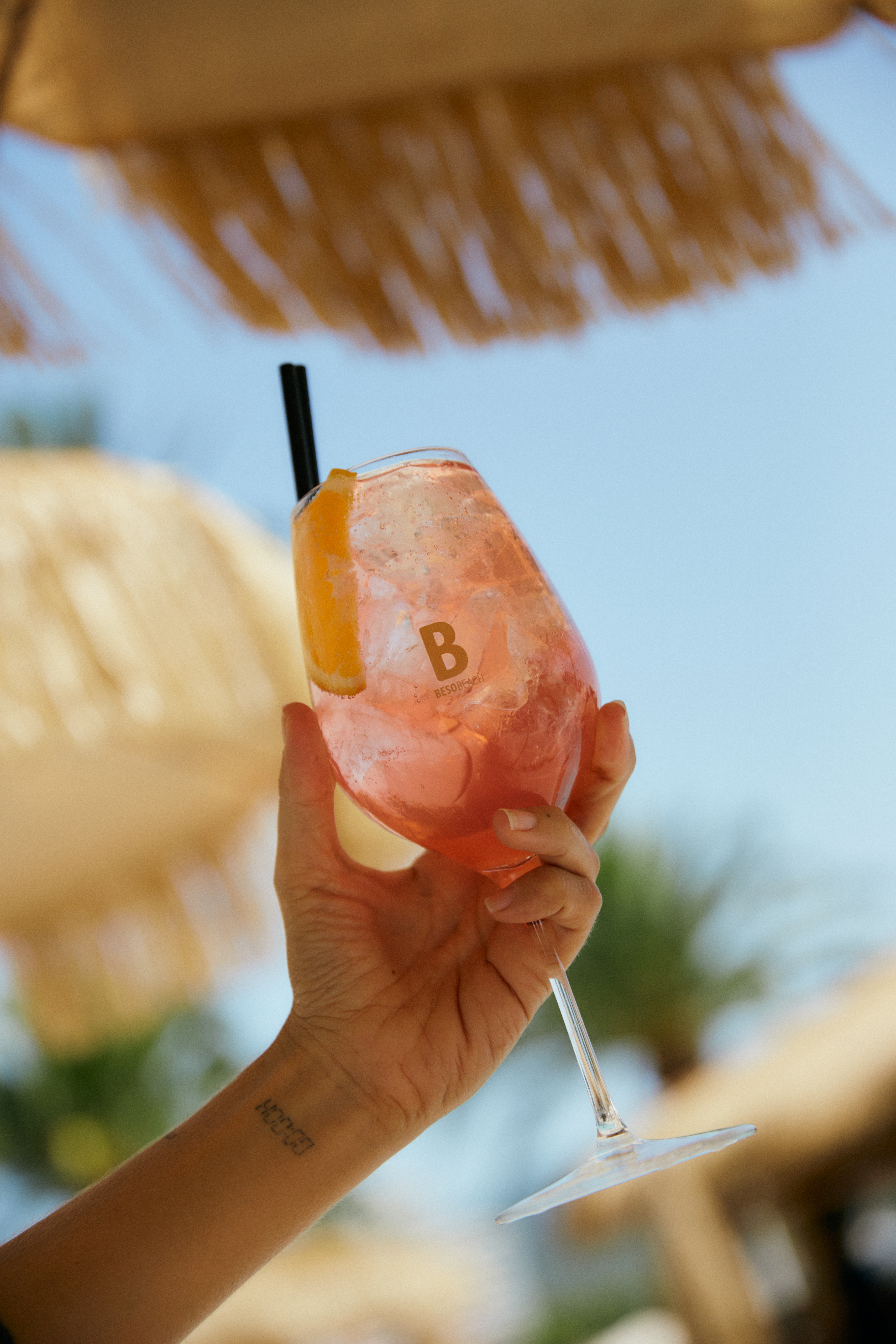 Hand holding pink Beso Beach cocktail with orange slice, straw, beach umbrella.
