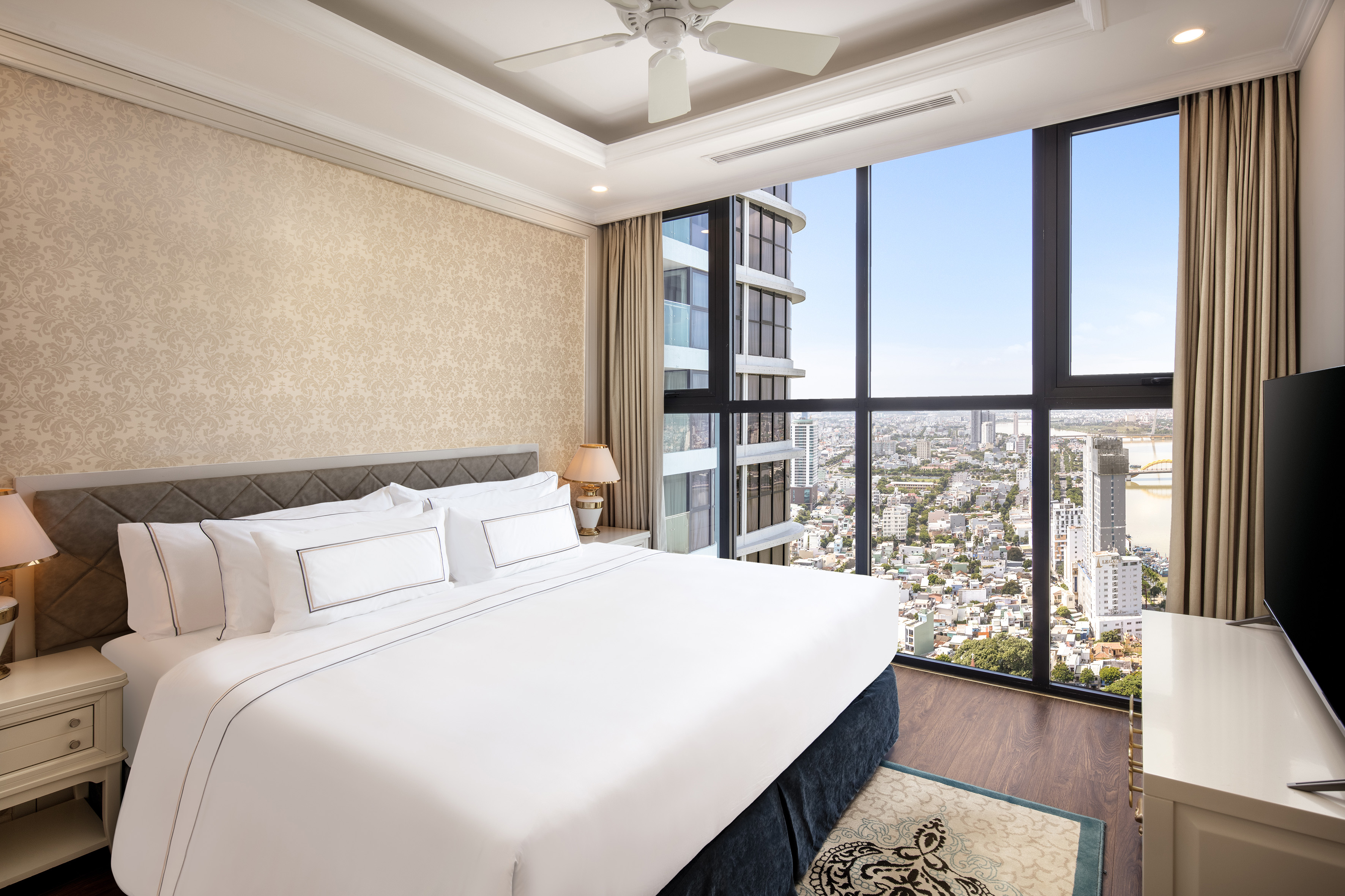 Luxury bedroom with white bed, panoramic city view, ceiling fan, and elegant decor.